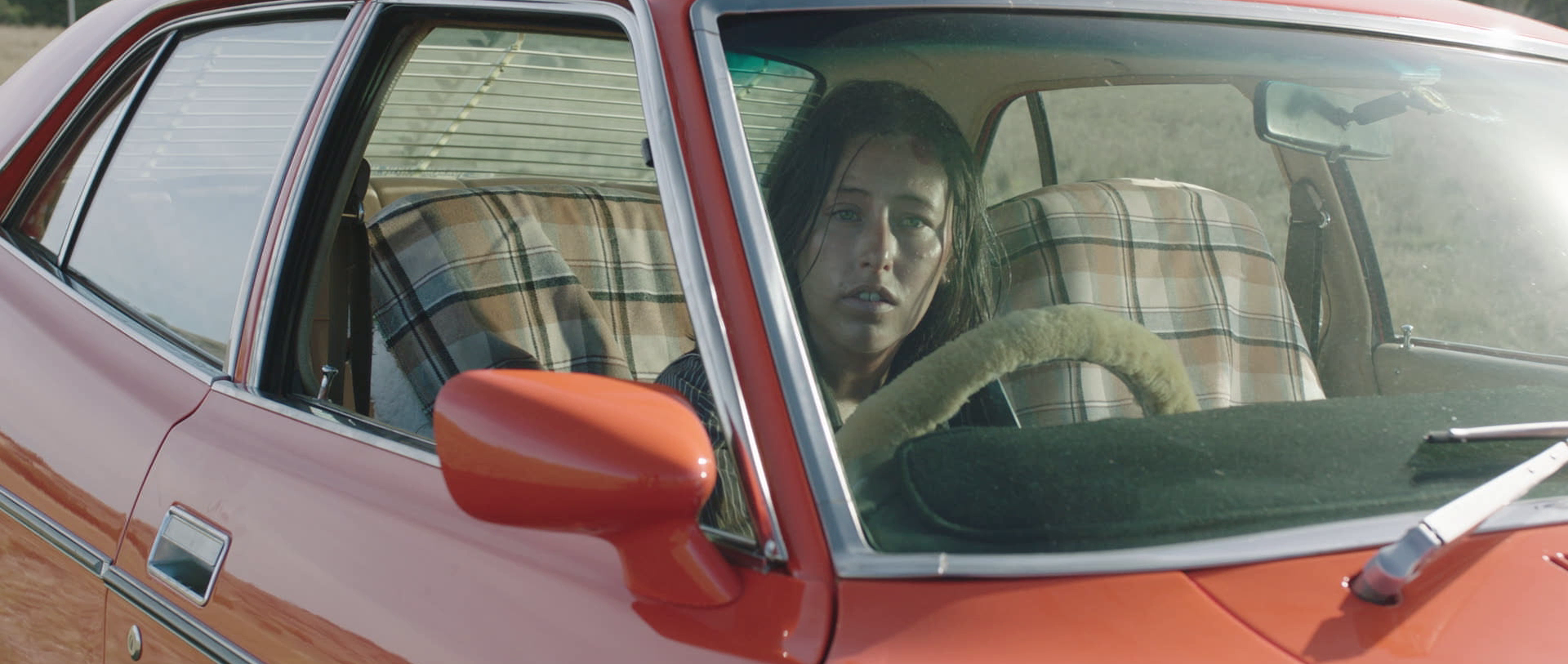 A woman with wet hair sitting inside a red vintage car with plaid-covered seats, looking out the driver's side window with a serious expression.