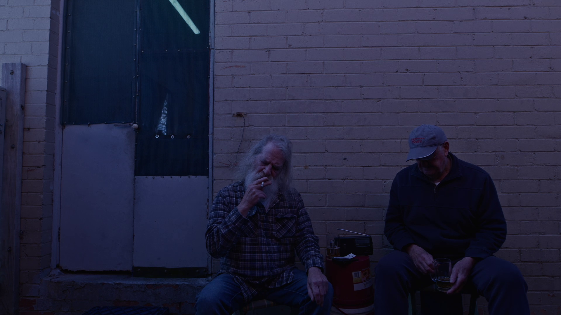 Two older men sitting outside against a brick wall, one smoking a cigarette and the other holding a glass of beverage.