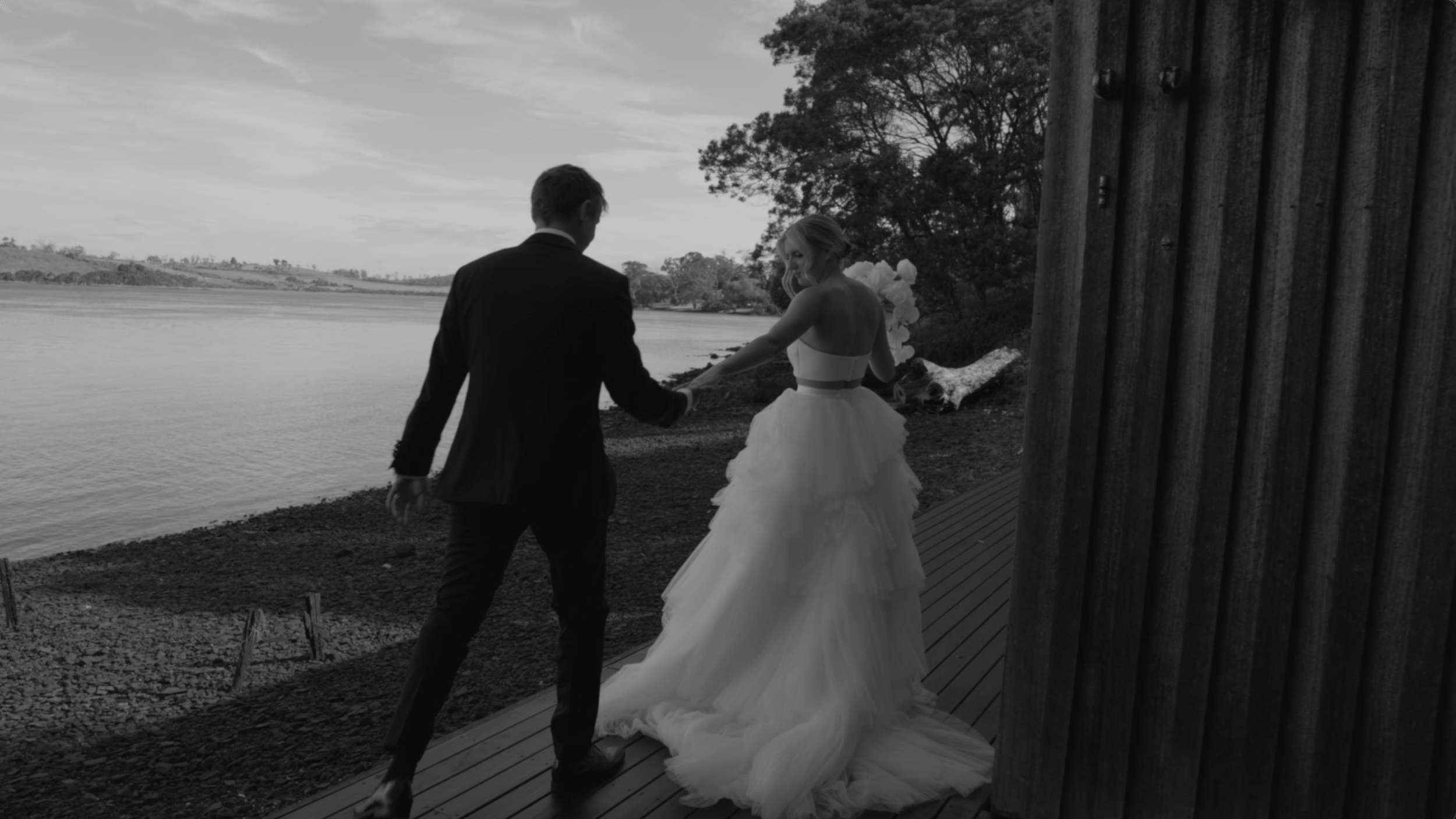 A bride and groom holding hands by a lake at sunset, with trees in the background, seen from behind.