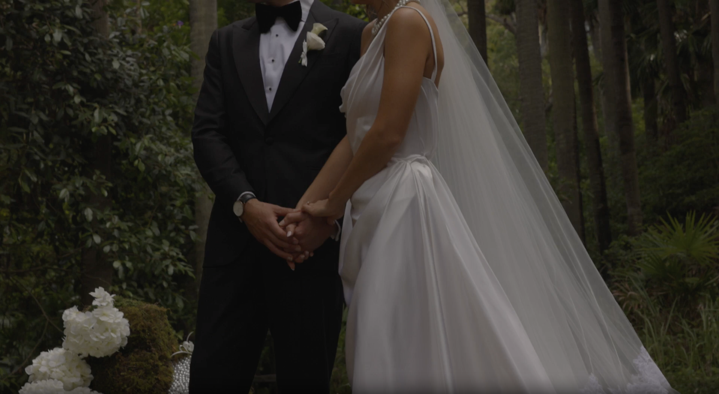 A bride and groom holding hands during their outdoor wedding in a wooded area. The groom is wearing a black tuxedo and the bride is in a white wedding dress with a long veil.