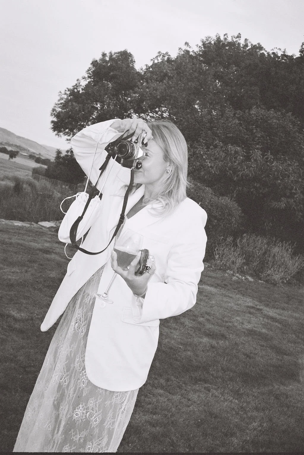 A woman in a white blazer and lace dress taking a photo outdoors with a digital camera, holding a wine glass and a phone in her other hand, standing on a grassy area with trees and hills in the background.