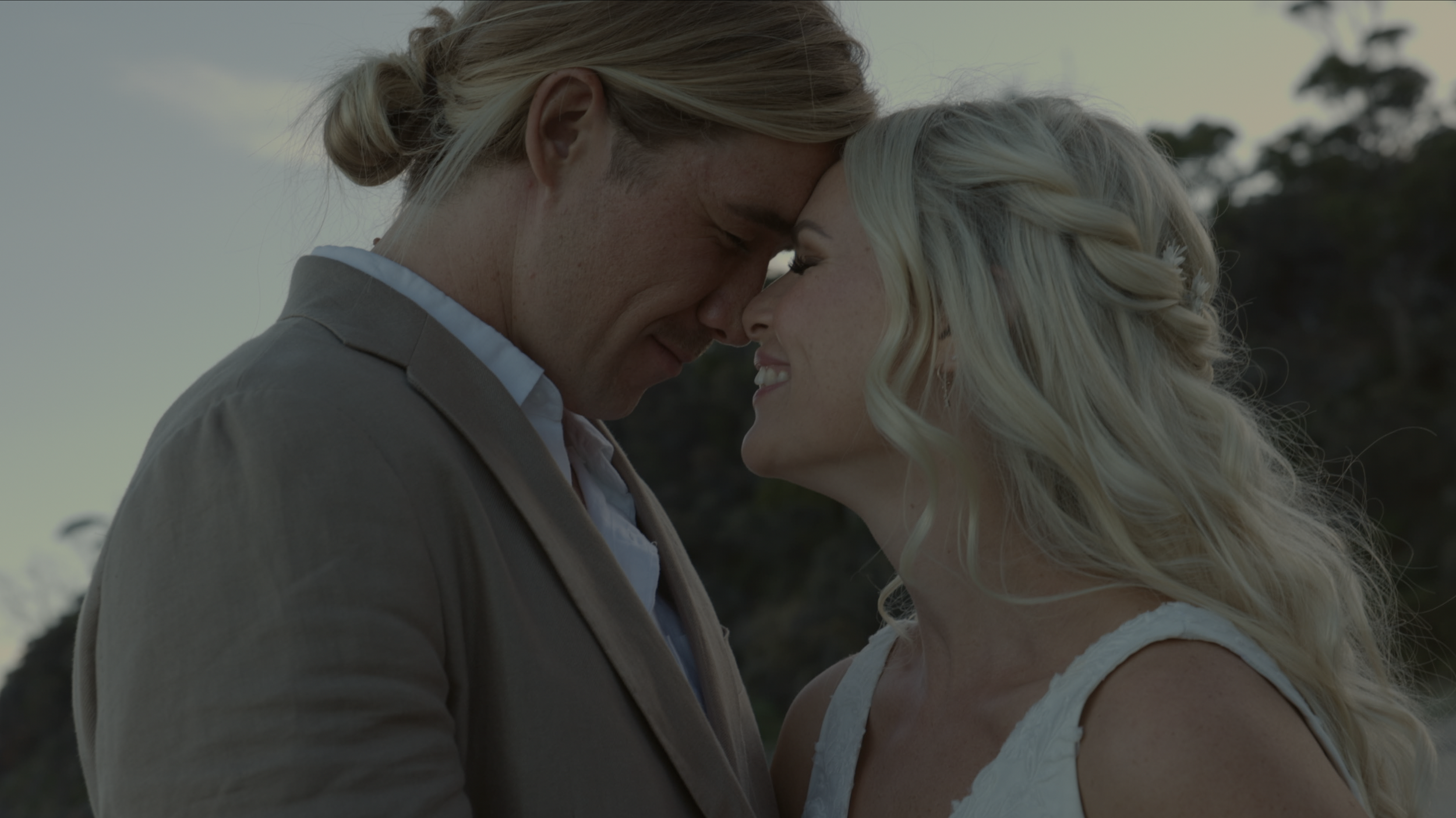 A close-up of a man and woman touching foreheads with their eyes closed, smiling softly against a natural outdoor background.