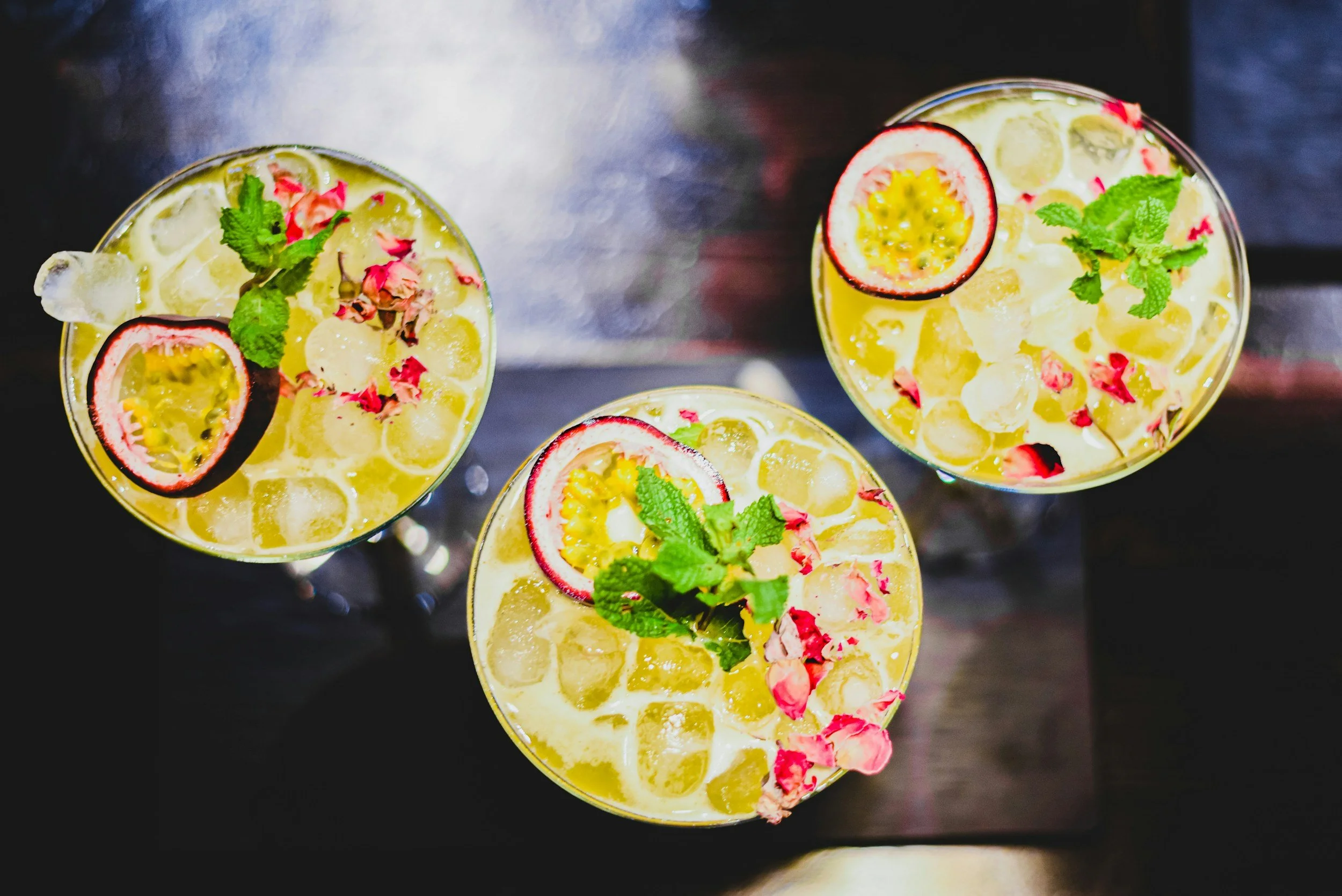 Three glasses of yellow cocktail garnished with passion fruit halves, mint leaves, and pink flower petals on ice cubes, viewed from above.