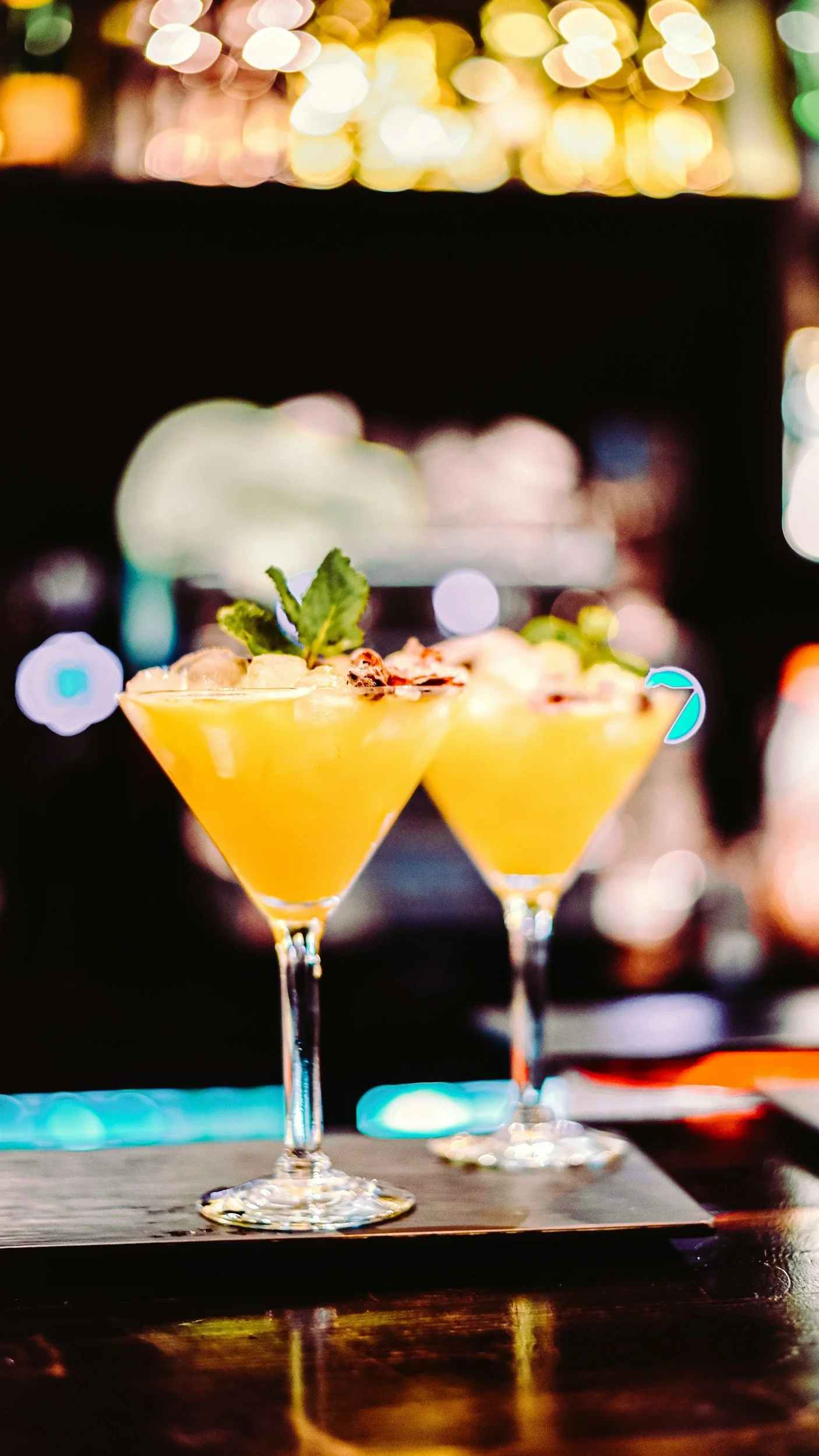 Close-up of two yellow cocktails in martini glasses garnished with mint leaves and flower petals, on a wooden surface with blurred colorful bokeh lights in the background.
