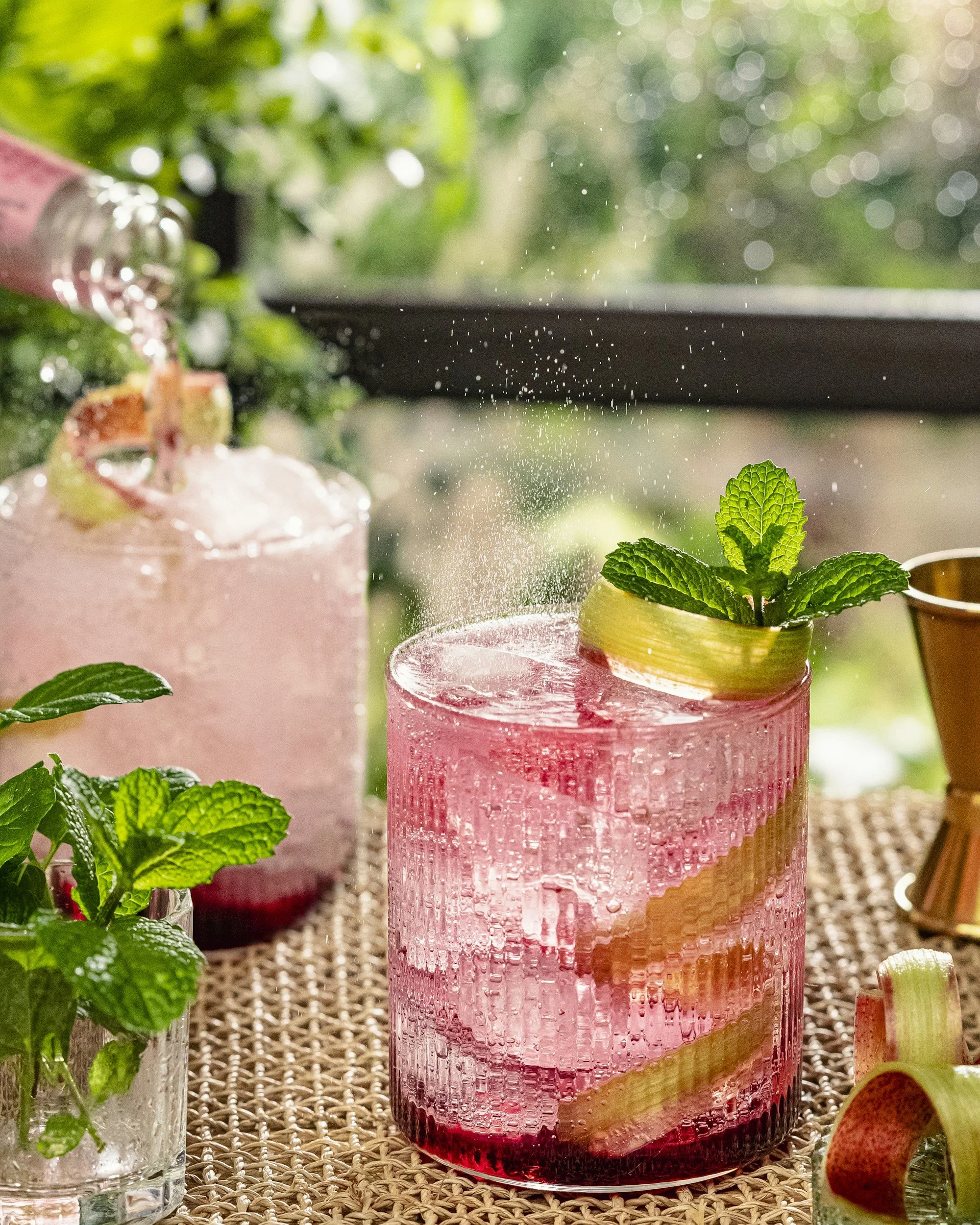 A pink cocktail garnished with a lemon peel and mint leaves, with a straw, on a woven mat with a blurry background of green foliage.