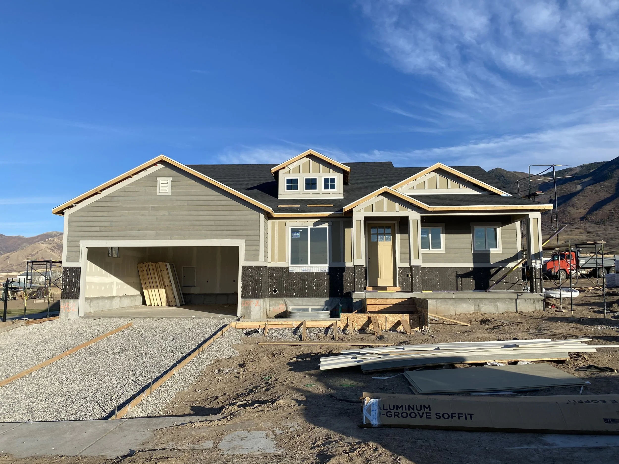 Under construction house with beige exterior, black roof, and gravel driveway, with mountains in the background and construction materials around.