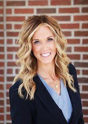 A woman with long, blonde, curly hair smiling against a brick wall background, wearing a dark blazer and light blue top.