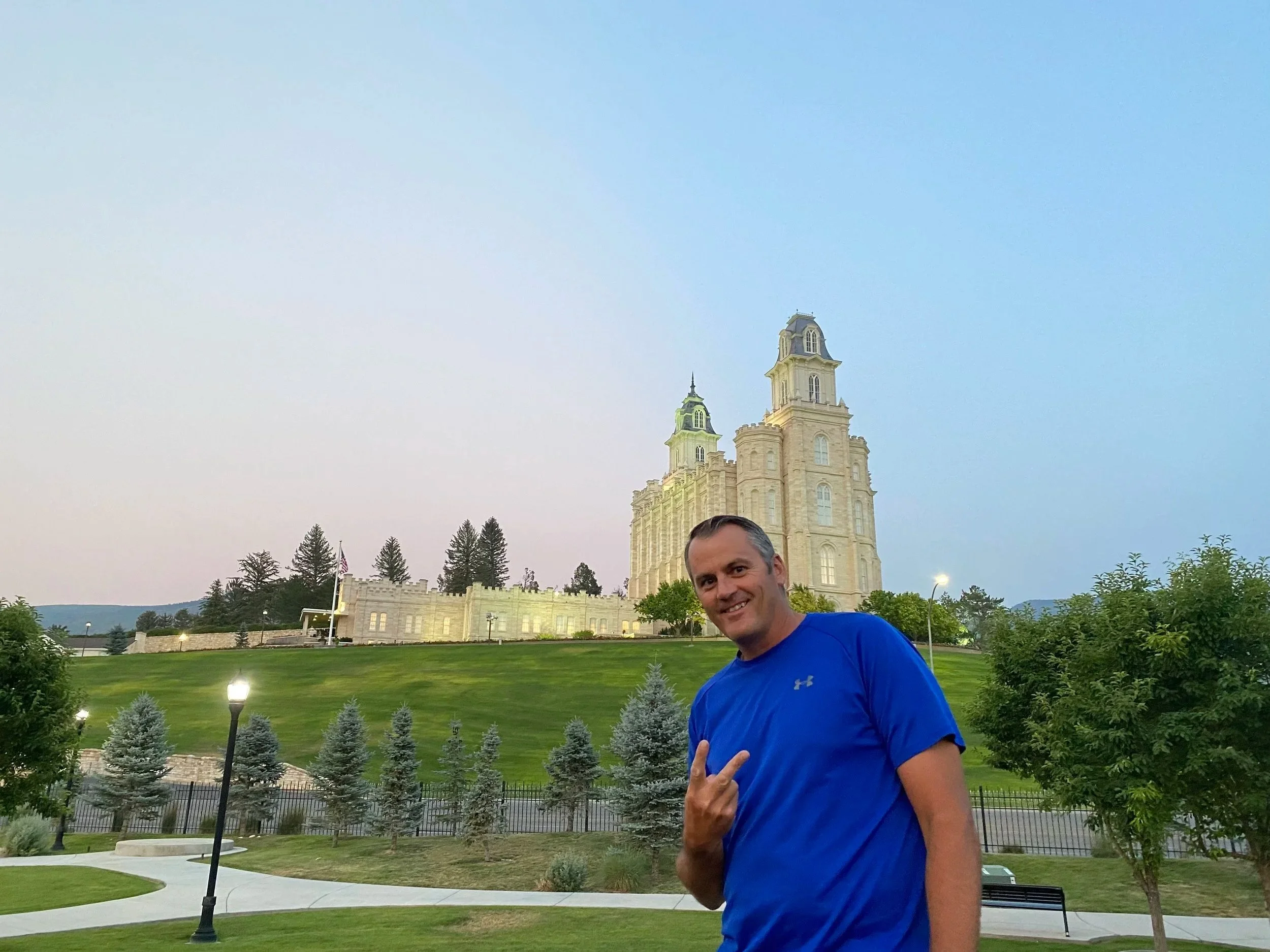Brandon Pehrson, general contractor and home builder at Manti Temple View Estates, in a blue athletic shirt making a peace sign next to a park with small trees, lampposts, and the Manti, Utah LDS temple in the background.