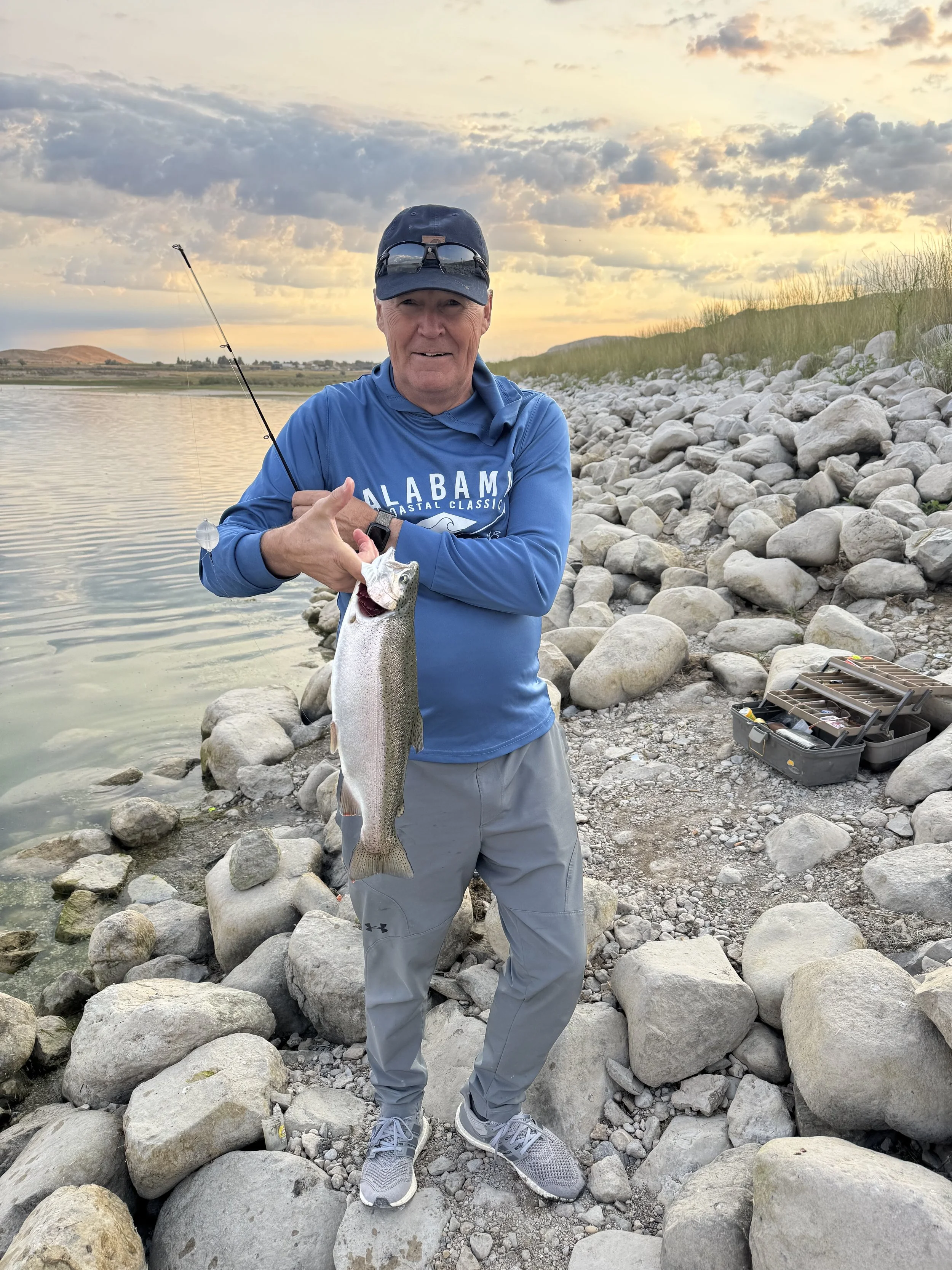 Chad Allred, developer of Manti Temple View Estates, stands on a rocky riverbank holding a freshly caught fish with a fishing rod in the background during sunset.