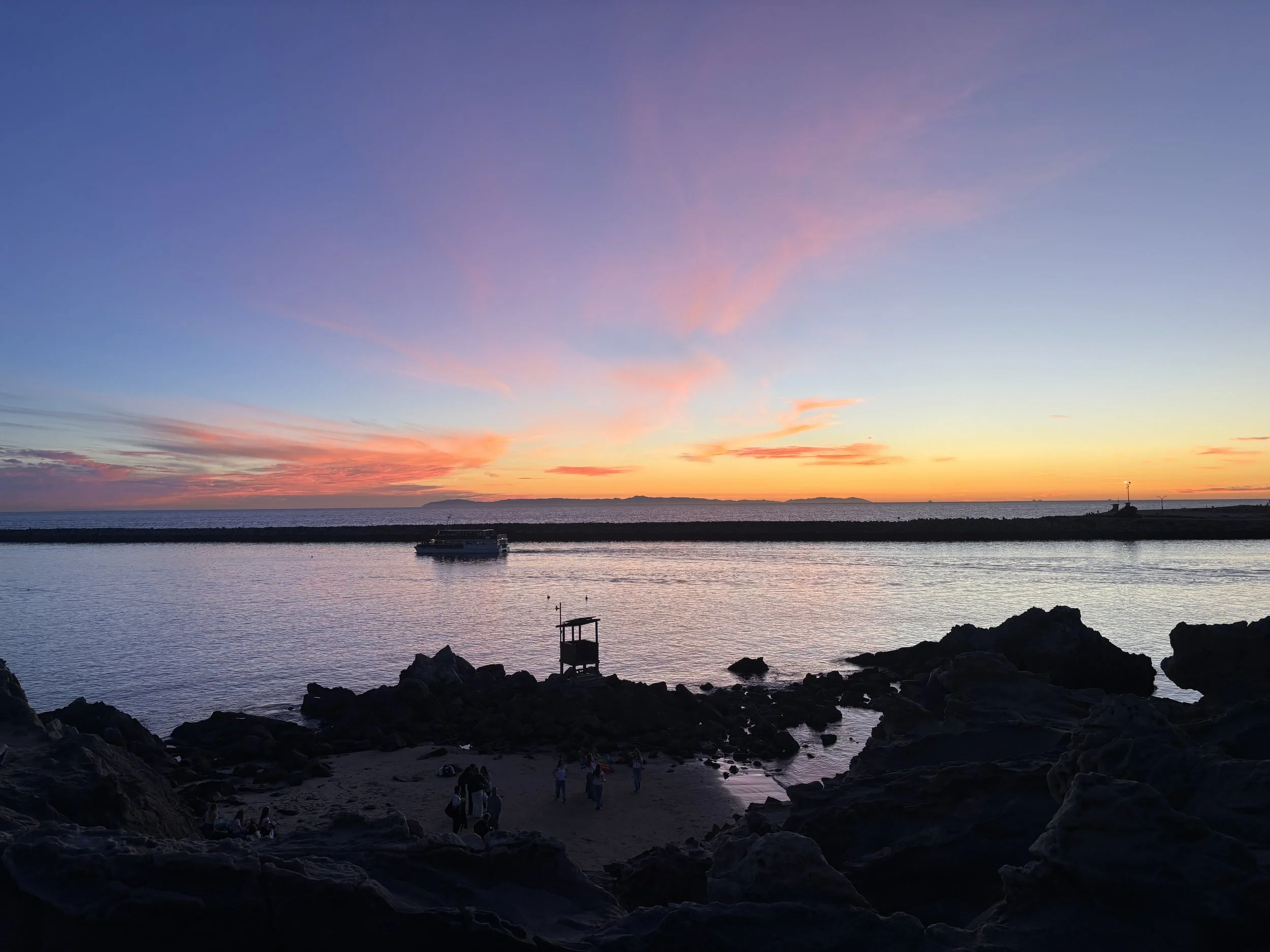 A sunset over the ocean with clouds in shades of pink, purple, and orange. Silhouetted rocks in the foreground with people near the shoreline and a boat in the water.