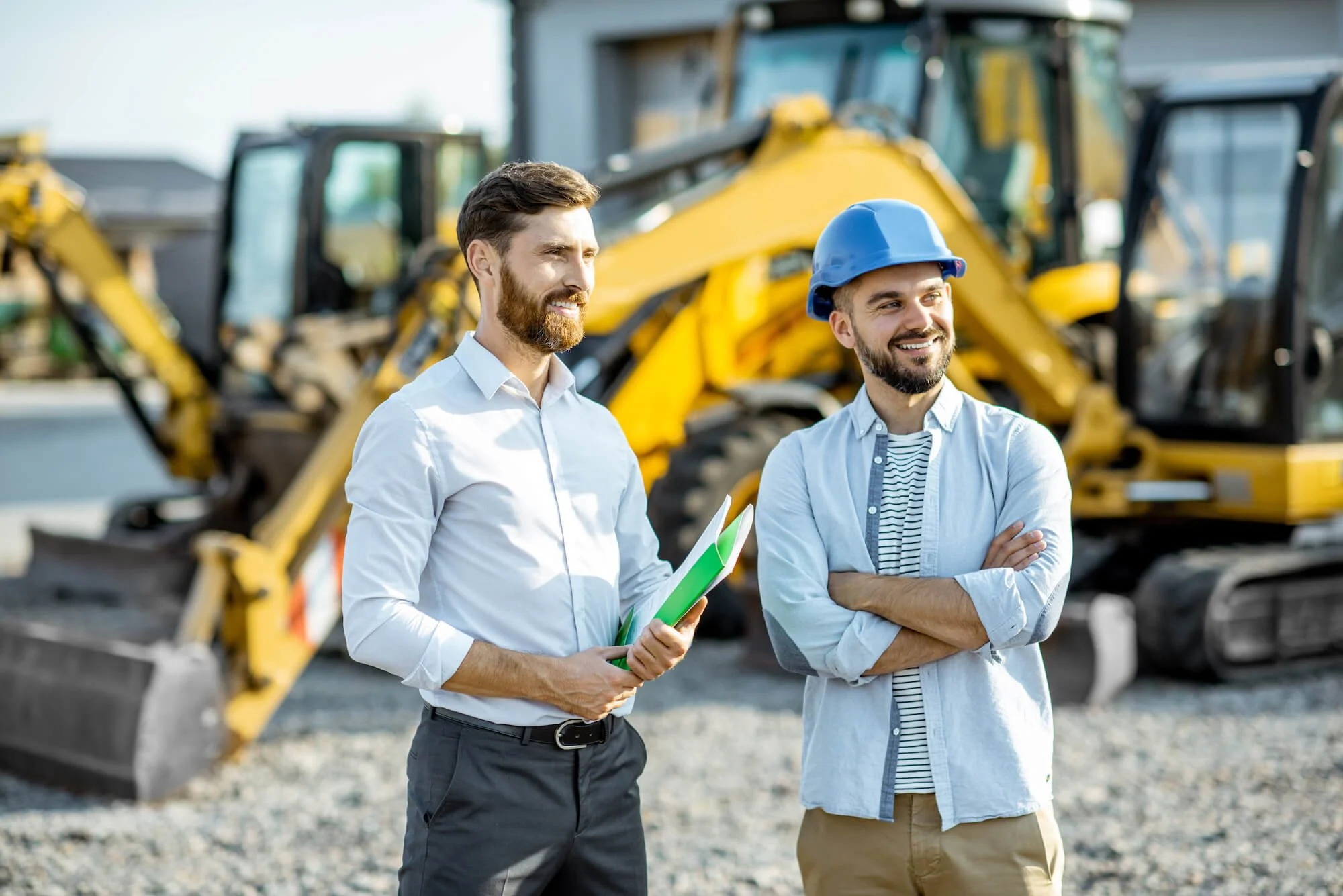 Two men standing outdoors near construction equipment, one holding a binder, both smiling, with excavators in the background.