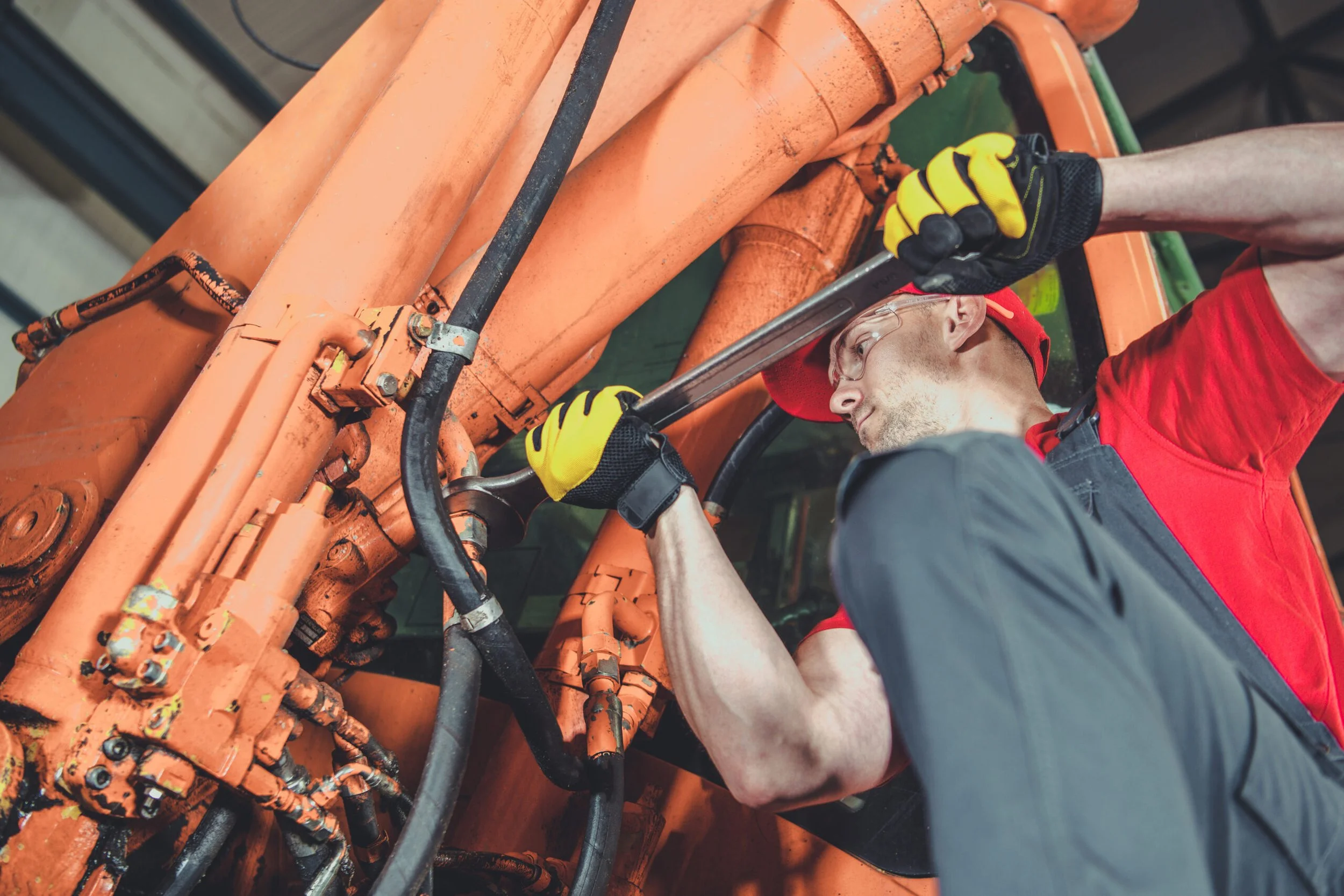 A man wearing glasses, a red shirt, and yellow gloves is repairing an orange heavy machinery with a large wrench.