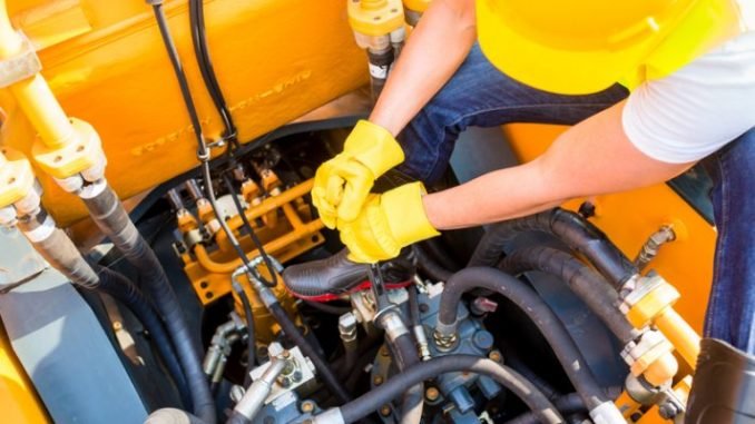A person wearing yellow gloves and a yellow hard hat working on a yellow industrial machine or vehicle engine with various pipes and hoses.
