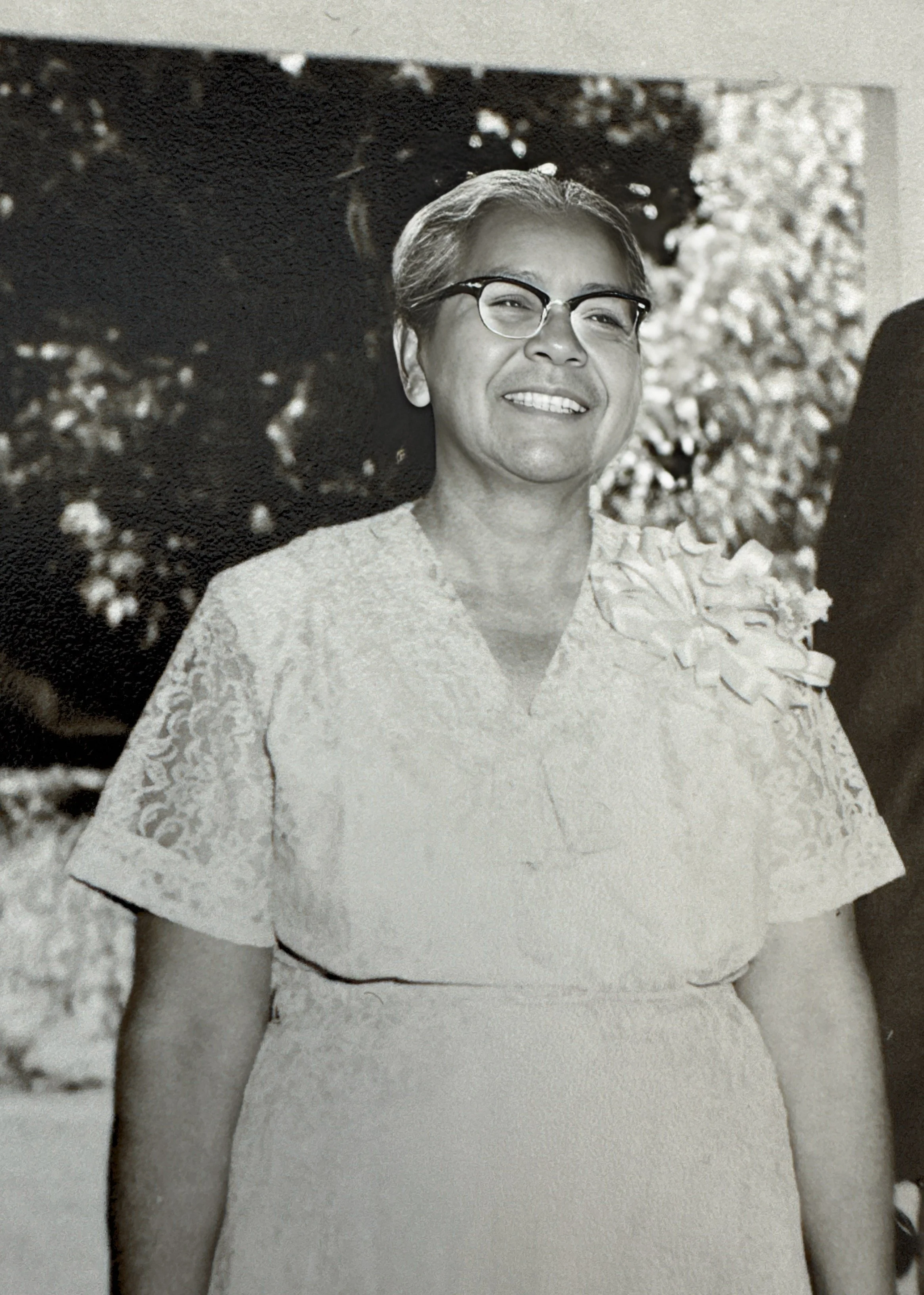 Black and white photograph of a woman wearing eyeglasses and a lace dress with a floral decoration on her shoulder, outdoors with trees in the background.