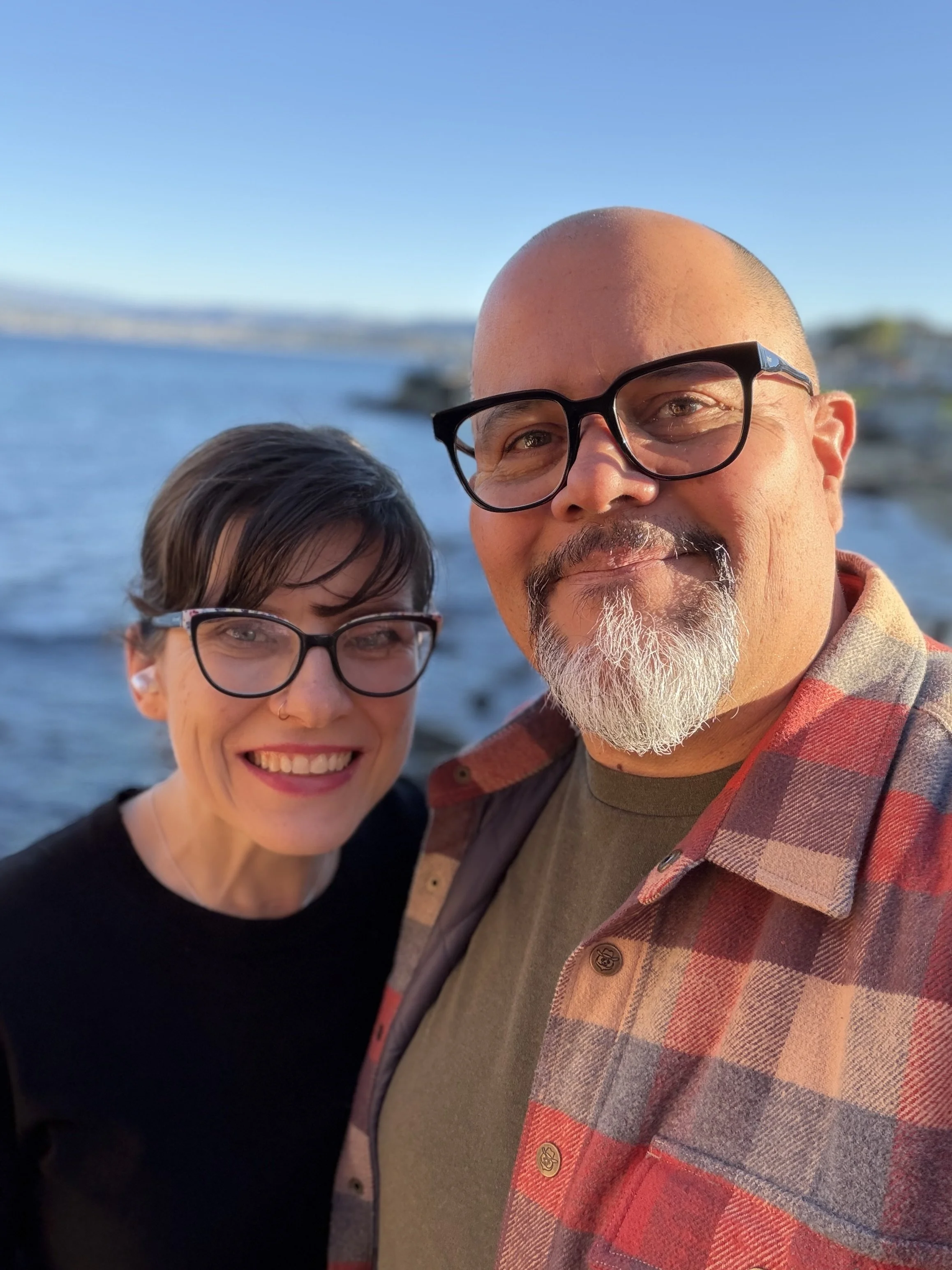 A smiling woman and a man with glasses taking a selfie outdoors near a body of water, with a clear blue sky in the background.