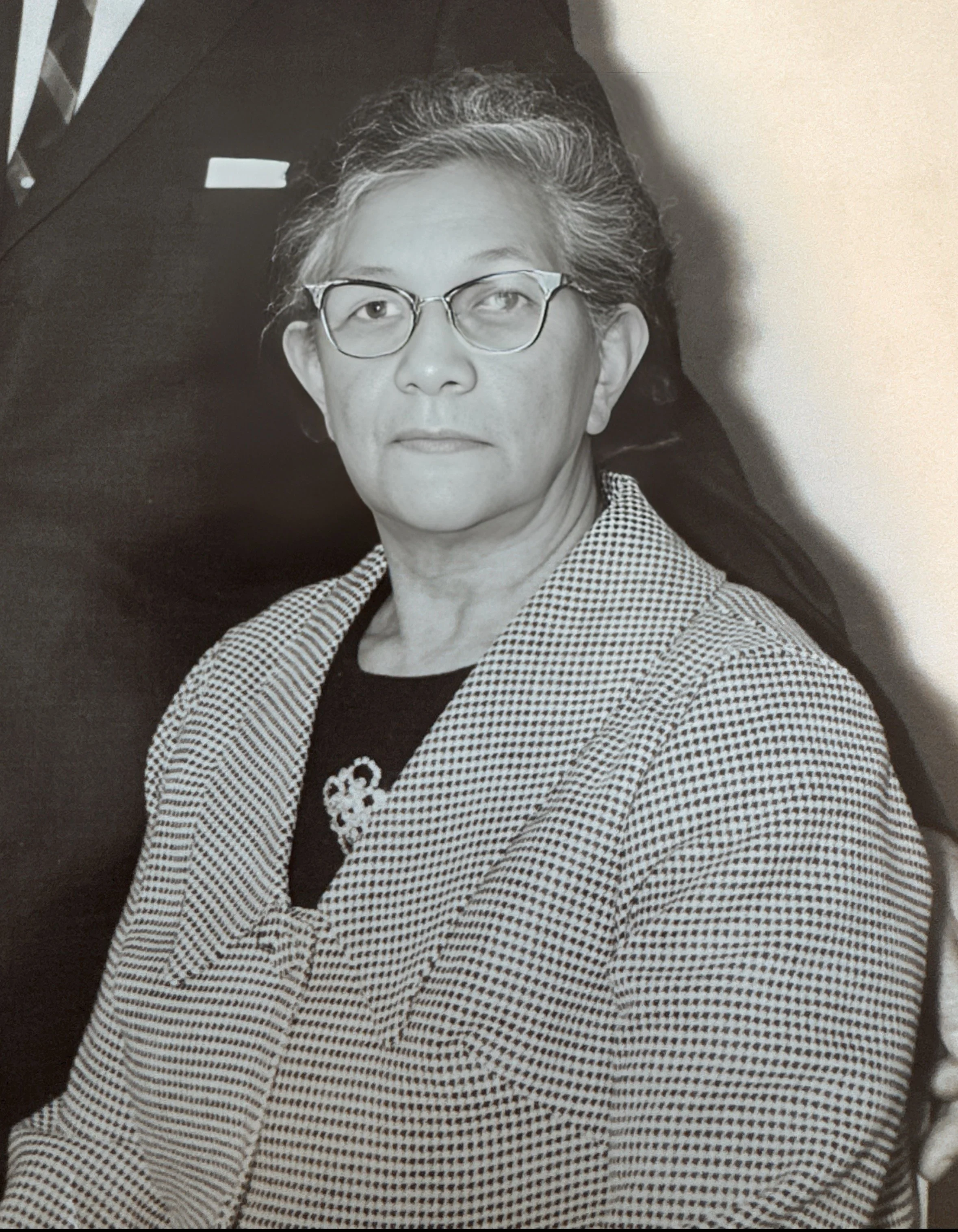 A black and white photo of a woman with short, gray hair, glasses, wearing a checkered blazer over a dark top, sitting in an indoor setting.