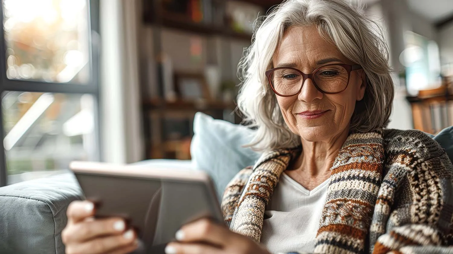 An elderly widowed woman with gray hair and glasses sits on a sofa, using a tablet. She is wearing a patterned sweater and appears calm the worry of finances is taken off her.