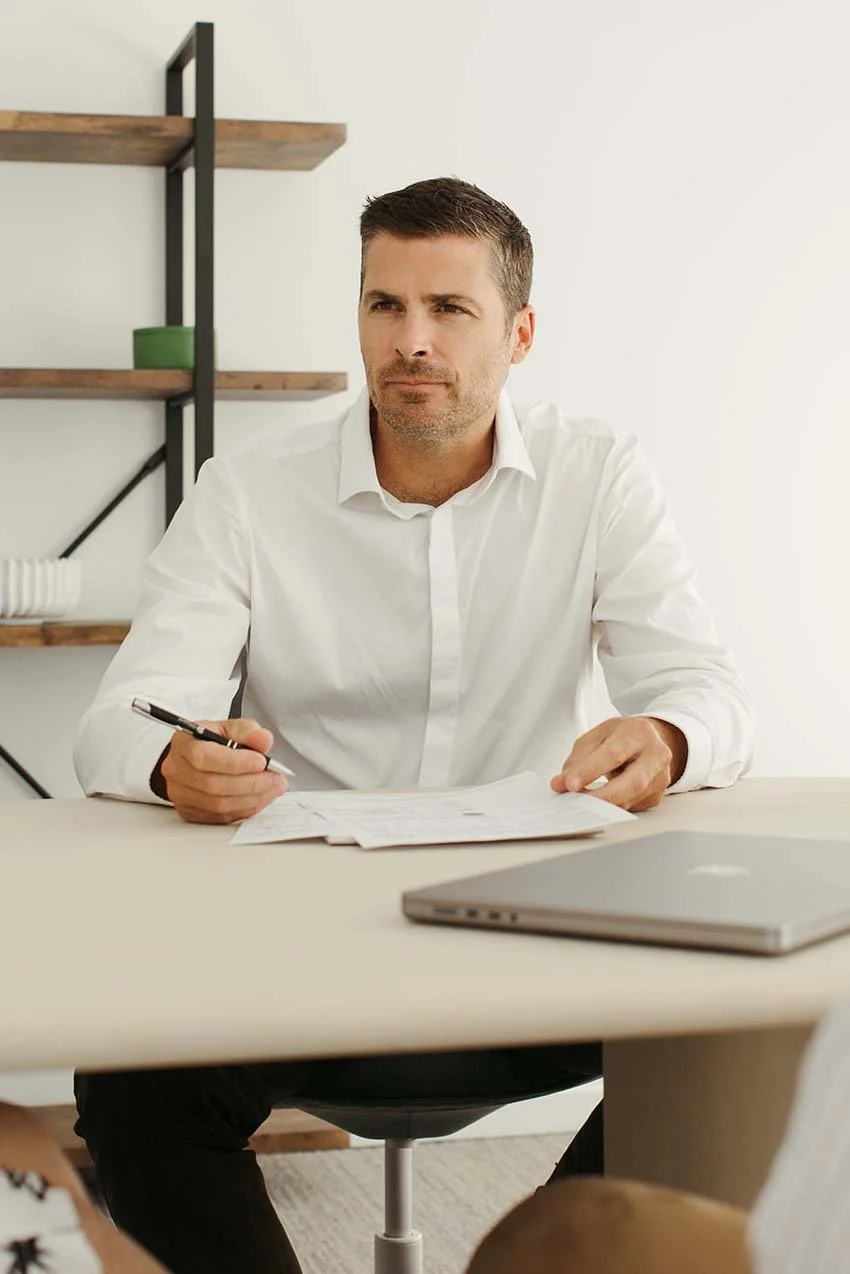 Steven Larman Financial Planner is in a white shirt sitting at a desk with papers and a laptop, holding a pen, in a modern office setting.