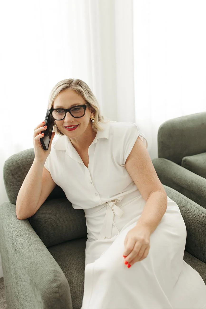 Andrea Paynter Financial Planner with blonde hair, black glasses, and red lipstick sitting on a green sofa, talking on a cell phone in a bright room with white curtains.