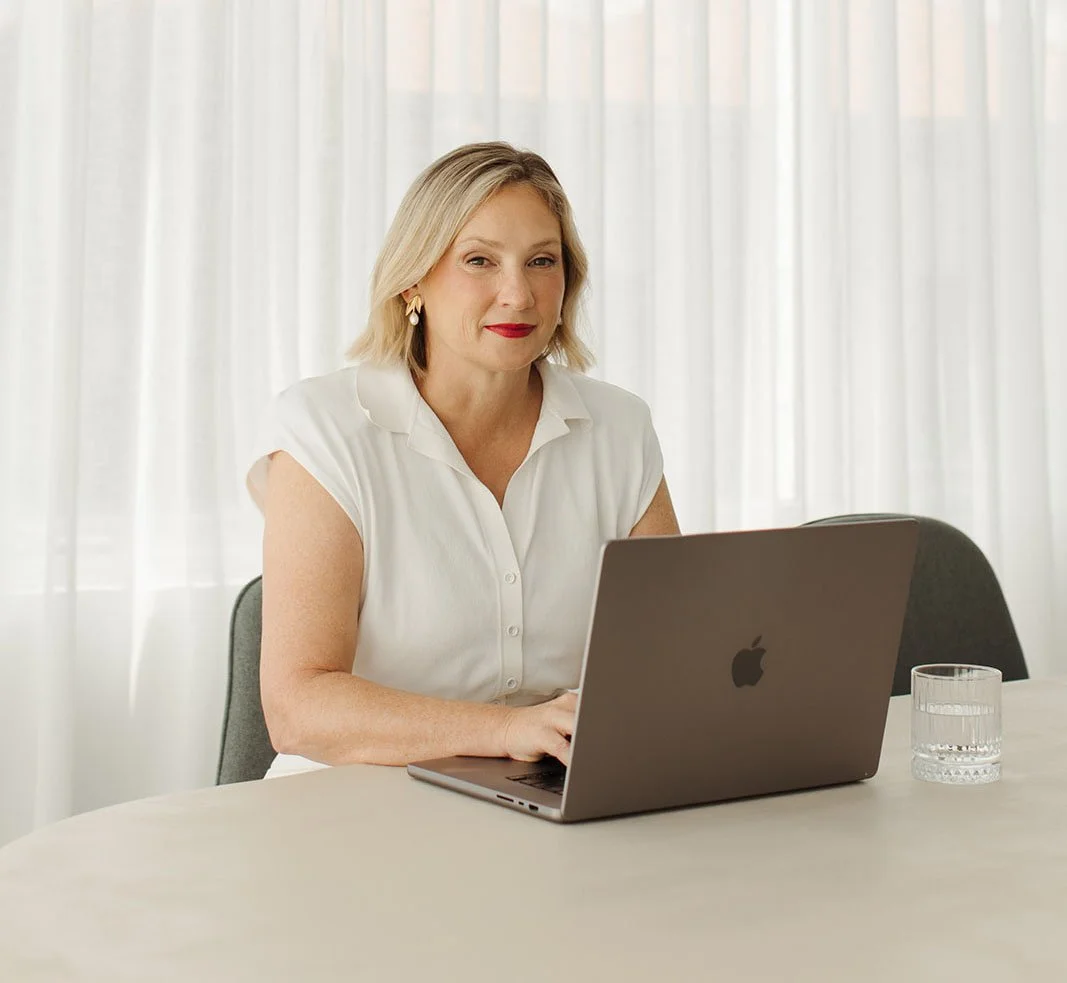 Andrea Paynter with blonde hair sitting at a table, working at financial planning on a silver MacBook laptop, with a glass of water nearby, in a bright room with white curtains.