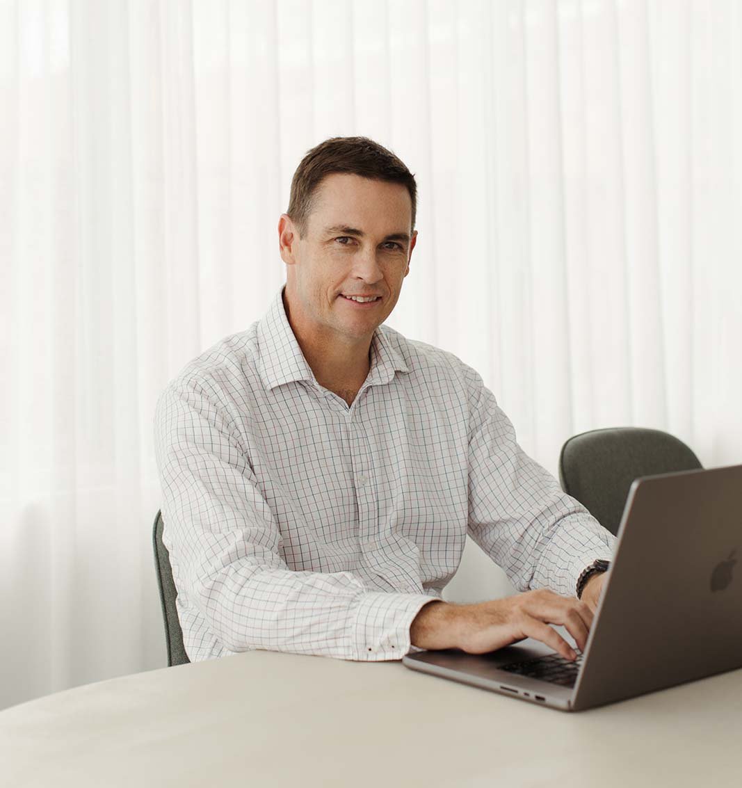 David Larman in a white checkered dress shirt sitting at a table with a silver laptop in front of him. He is smiling to this Financial Planning clients in his sunny Newstead office.