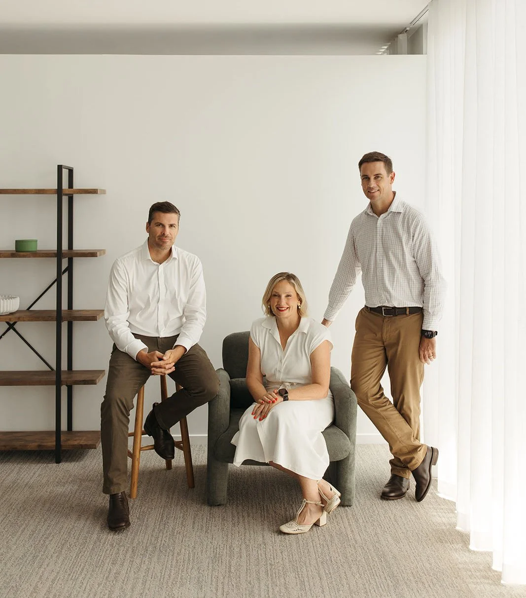 The team at Bailiwick in an office setting smiling and posing for a photo. Andrea Paynter is sitting in a gray chair, and David and Steve Larman are next to her.