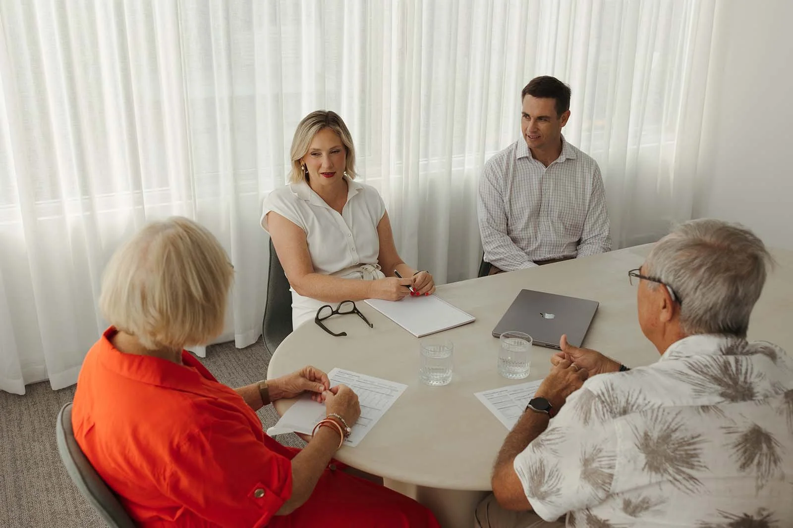 Anrread Paynter and David Larman sitting at a round table having a discussion with an older couple about financial planning. The table has laptops, papers, and glasses of water on the table.