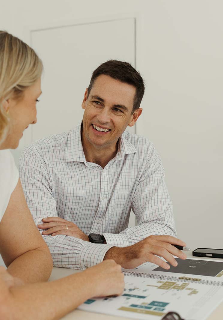 David Larman is  sitting at a table having a conversation with Andrea Paynter, with papers and documents in front of them. They are discussing financial planning for a married 50's couple pre-retiree stage.