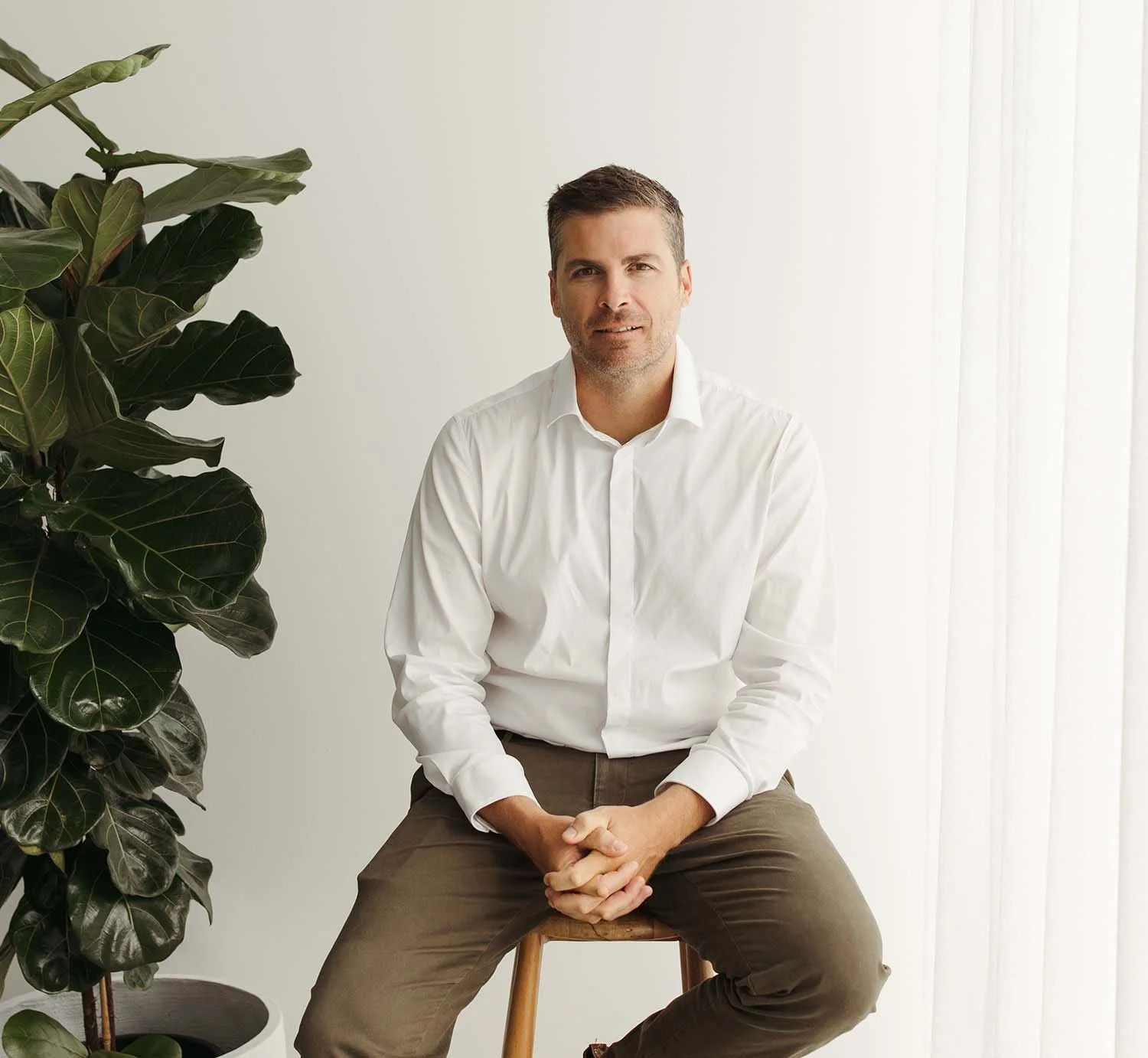Steven Larman, Financial Planner at Bailiwick Financial Planning, sits on a wooden stool near a large leafy plant in his bright Newstead office.