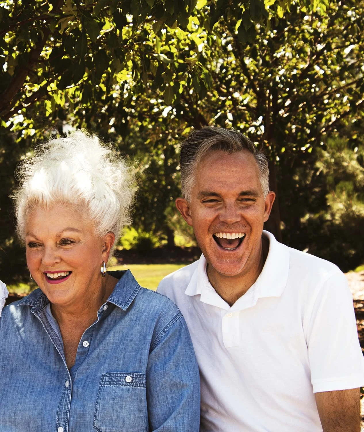 A smiling elderly woman with white hair and a man with short grey hair, both sitting outdoors under a tree.