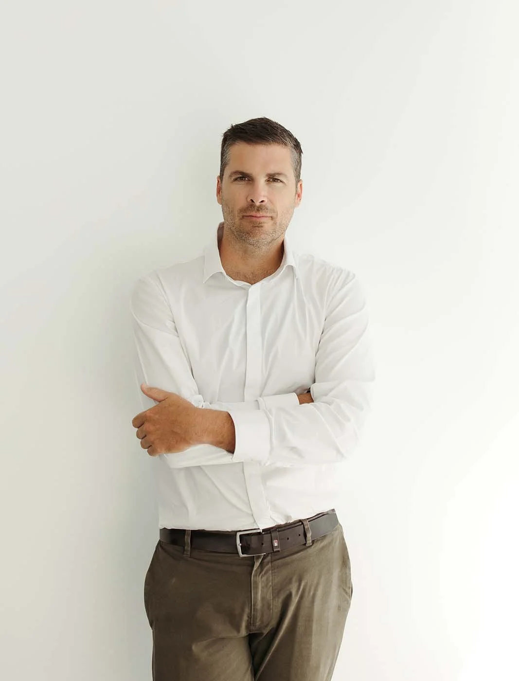 Steven Larman Financial Planner with short dark hair and light stubble, wearing a white button-up shirt and khaki pants, stands against a plain white wall in his Newstead office. He is looking directly at the camera.