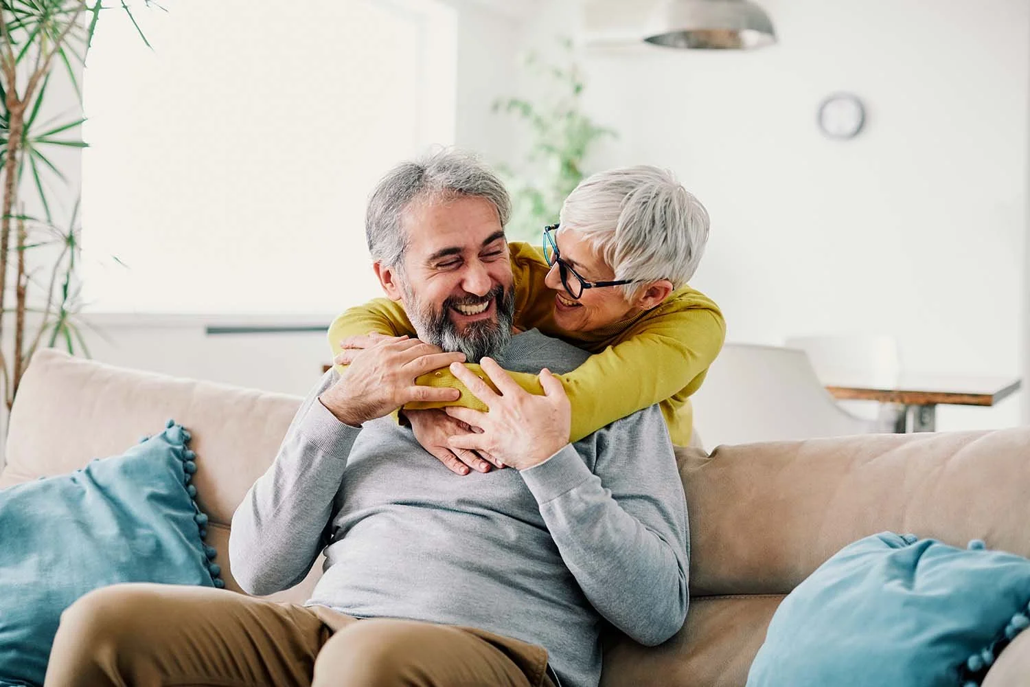 An older man sitting on a sofa with his retired wife hugging him from behind, both smiling and enjoying retirement.