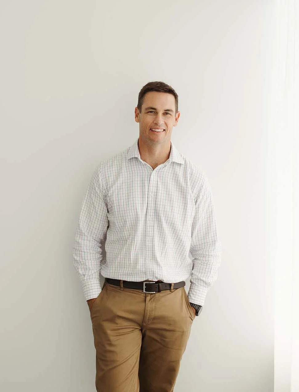 David Larman Financial Planner is in a white checkered shirt and khaki pants standing against a plain white wall, smiling with hands in pockets.