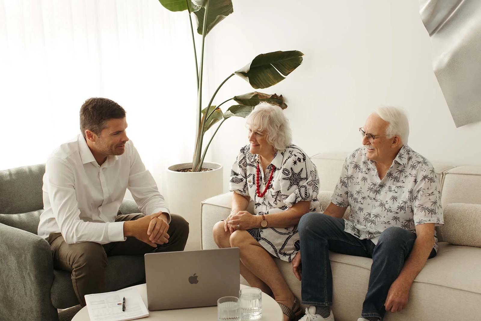 David Larman talking with an elderly retired couple in a bright office with a large potted plant, a MacBook on the coffee table, and glasses of water.