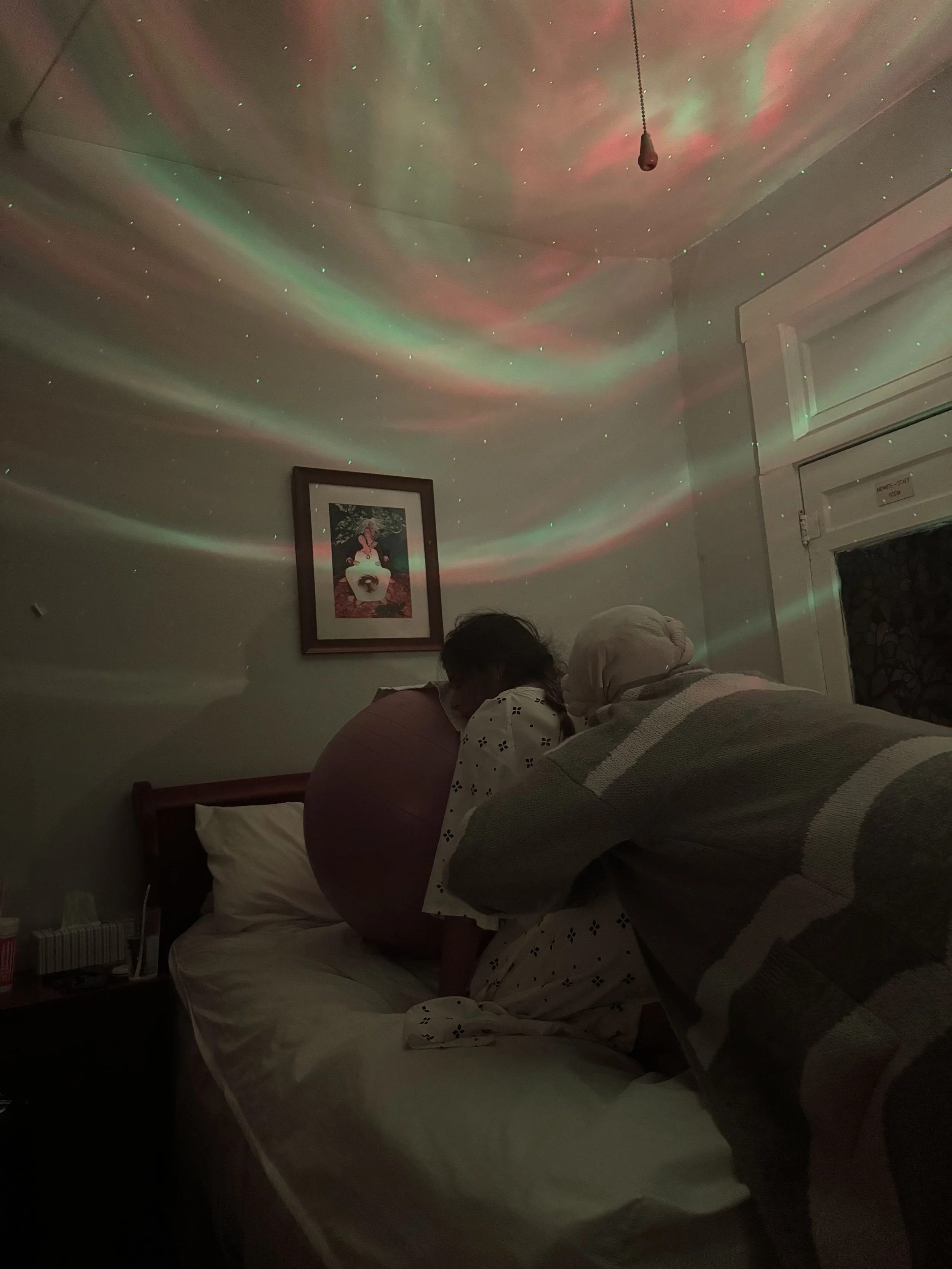 Two people are sitting on a bed in a dimly lit room with colorful lights projected onto the ceiling, creating a starry sky effect. One person is leaning forward while the other person, wearing a headscarf, is sitting beside them. The room has a framed picture on the wall and a nightstand with some items on it.