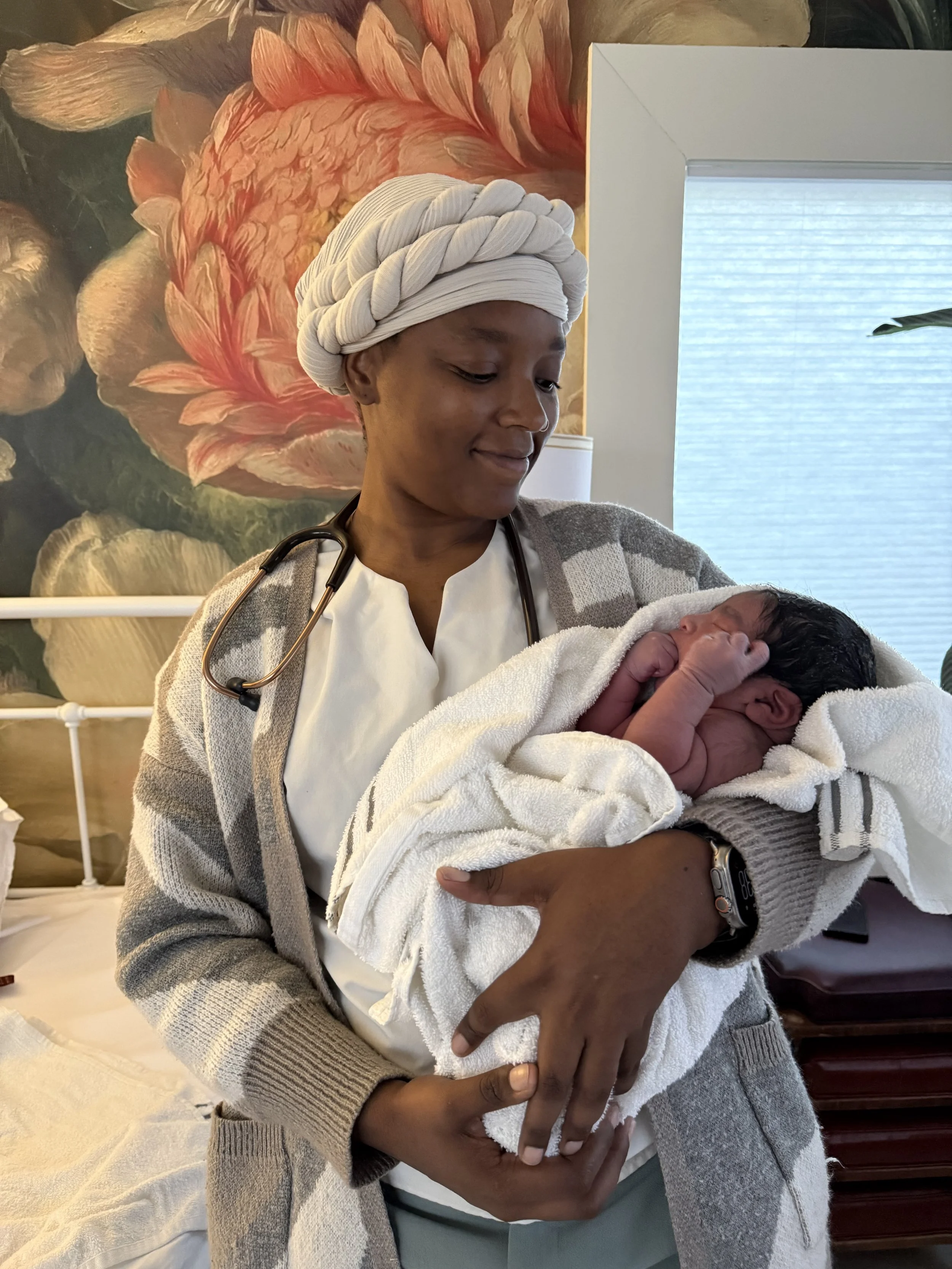A woman wearing a white headwrap, a gray cardigan, and a stethoscope around her neck is holding a newborn baby wrapped in a white towel, looking down at the baby with a gentle smile. The background features a large floral painting and a window.