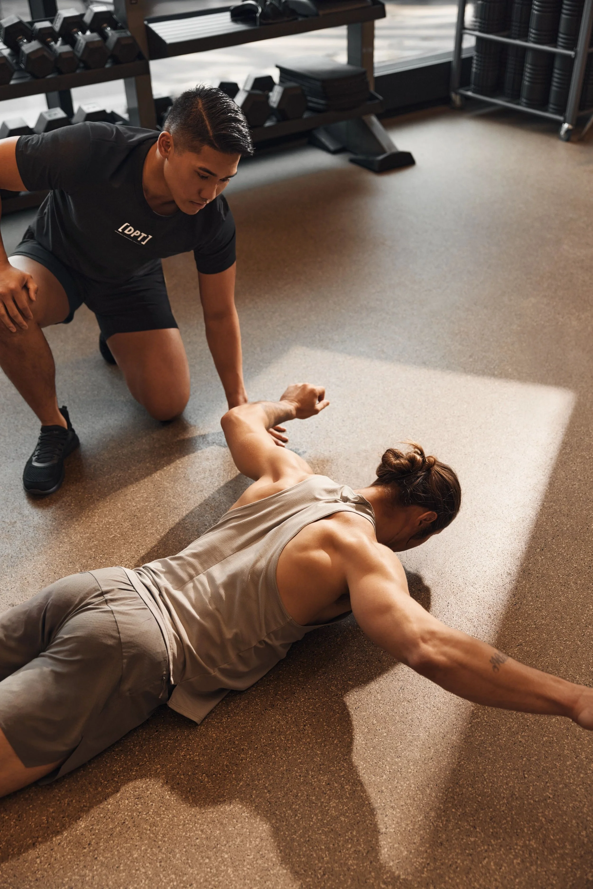 A trainer assisting a woman with stretching on the gym floor, with weights and exercise equipment in the background.