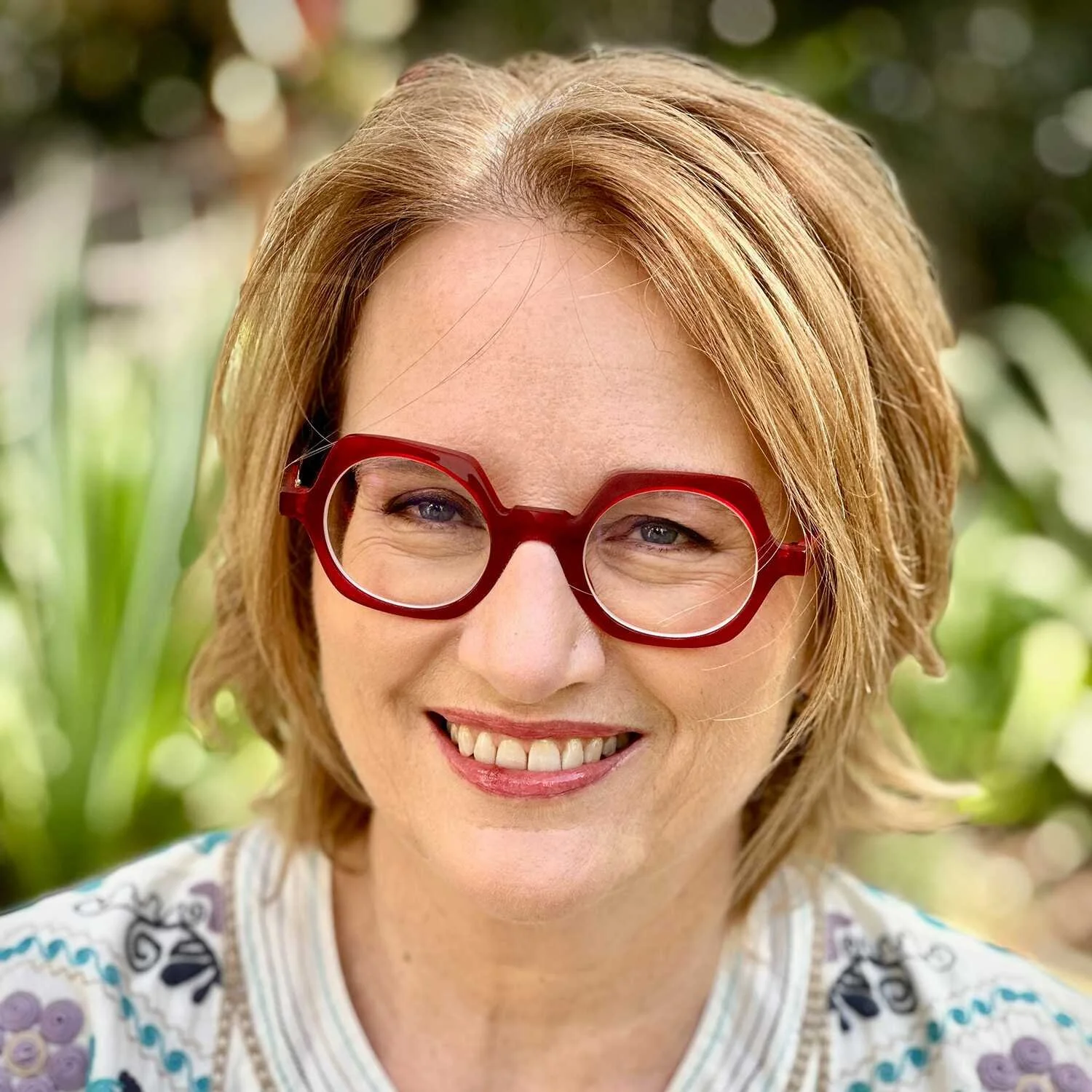 Writer Cynthia Banham with red hair and glasses smiling outdoors with greenery in the background.