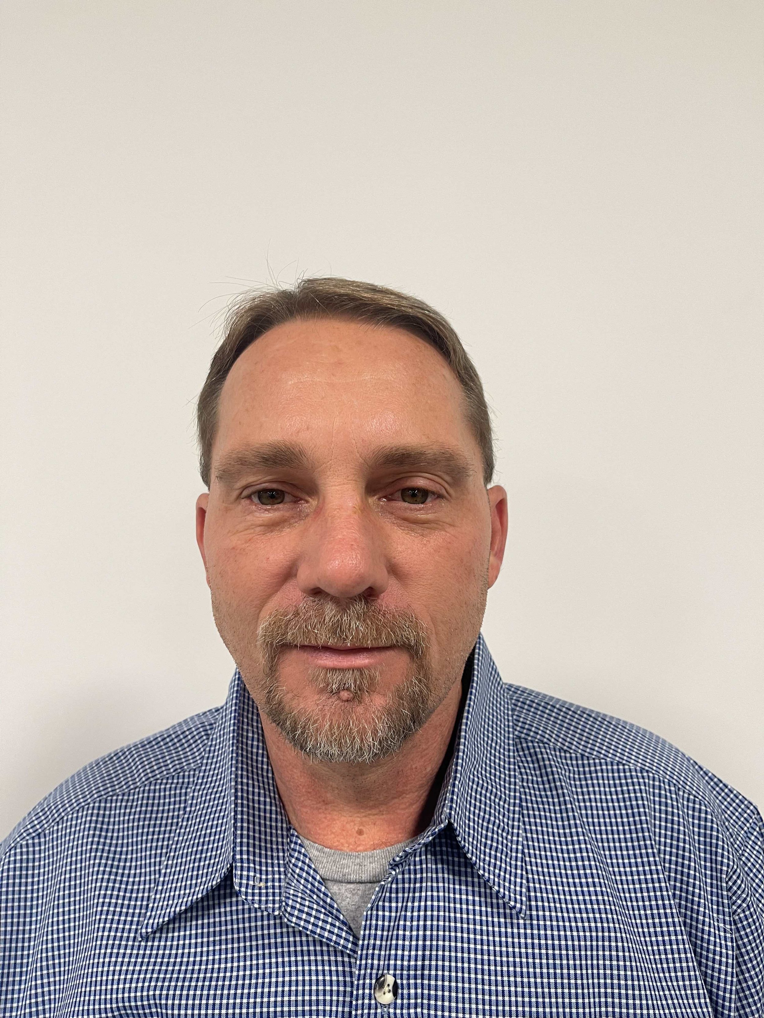 A middle-aged man with short brown hair, a goatee, and a mustache, wearing a blue checkered shirt, standing against a plain white wall.