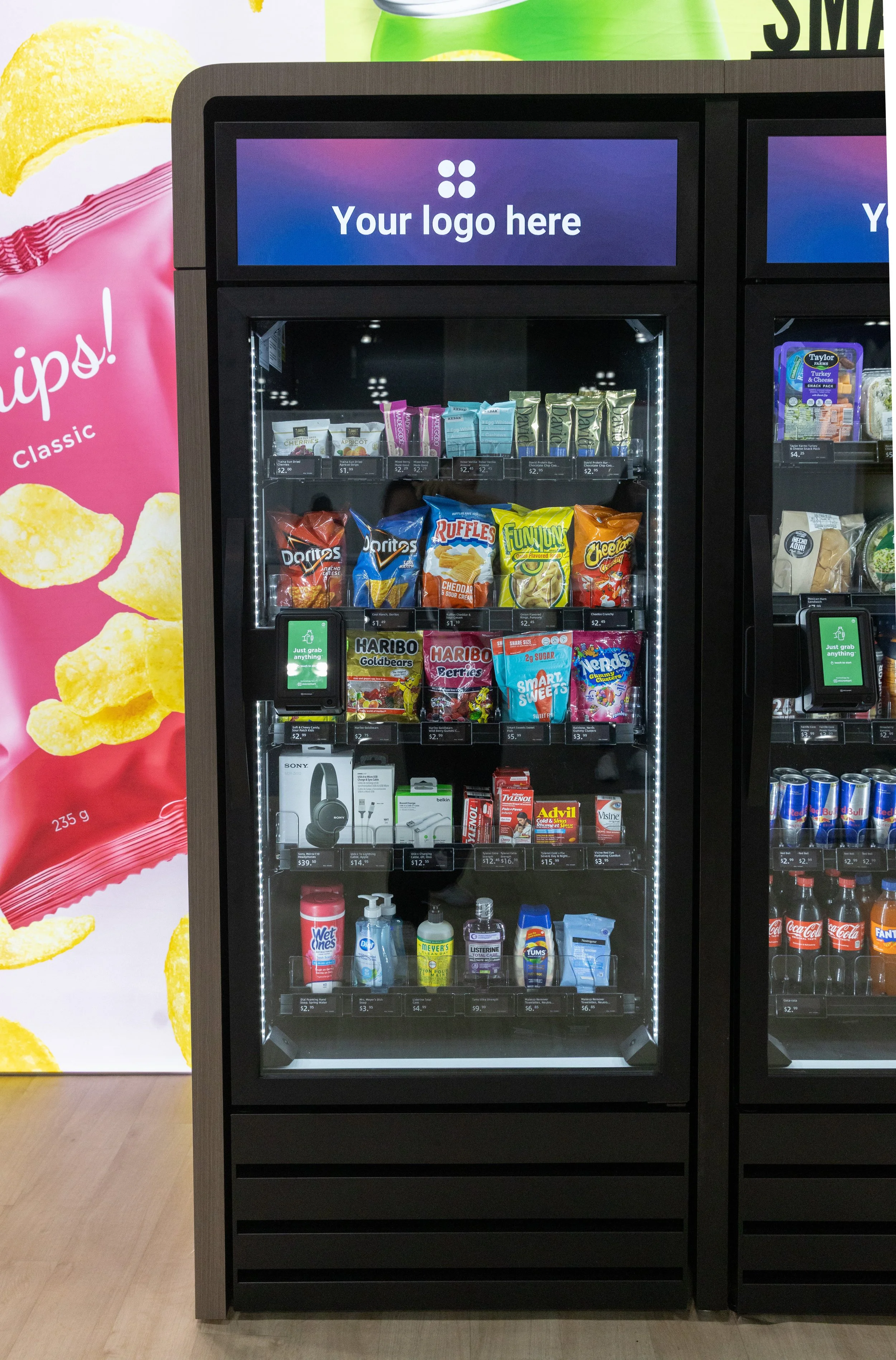 A smart market with snacks, drinks, and health products inside, with a digital screen above that says 'Your logo here', located next to a large advertisement for potato chips.