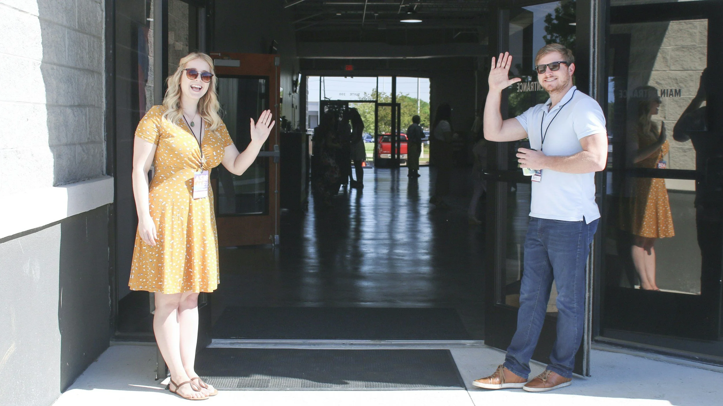 A young woman and a young man with sunglasses are standing outside an entrance, waving and smiling at the camera during a sunny day.