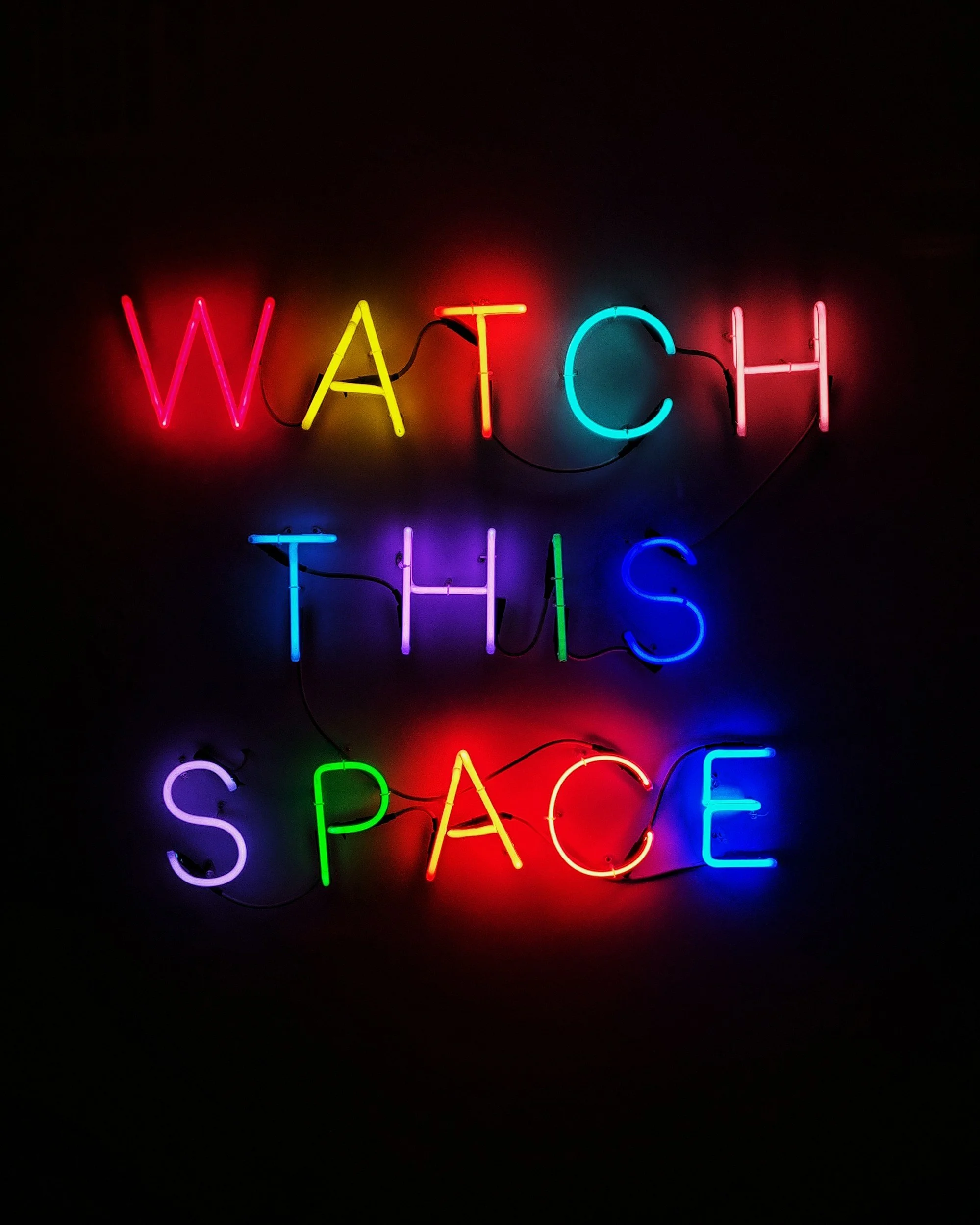 Neon sign saying "Watch this space" in colorful letters against a black background.