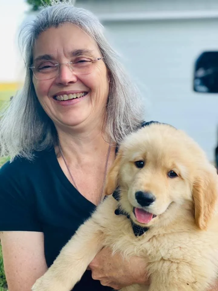 An elderly woman with gray hair and glasses smiling, holding a golden retriever puppy outdoors.