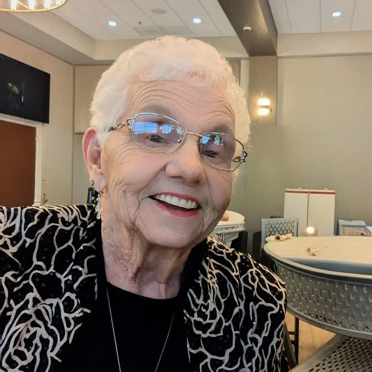 A smiling elderly woman with white hair, glasses, earrings, and a patterned black and white top, sitting in a restaurant or cafe.