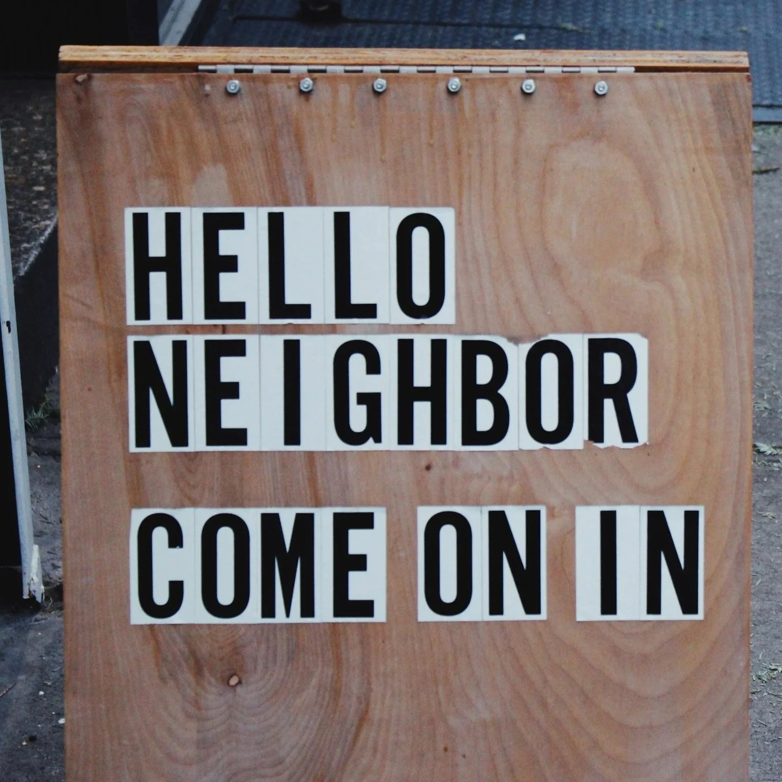 Signboard with a wooden background displaying the message 'Hello Neighbors, Come On In' in black letters on white blocks.