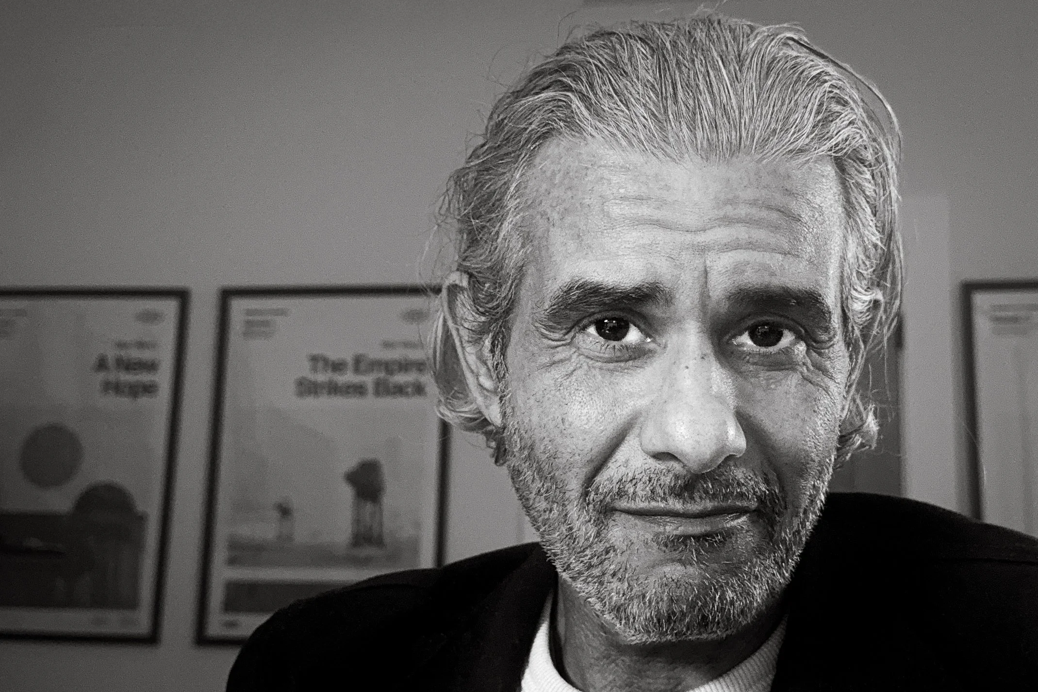 Black-and-white close-up portrait of Mo Husseini, smiling slightly, with framed posters on the wall behind him.