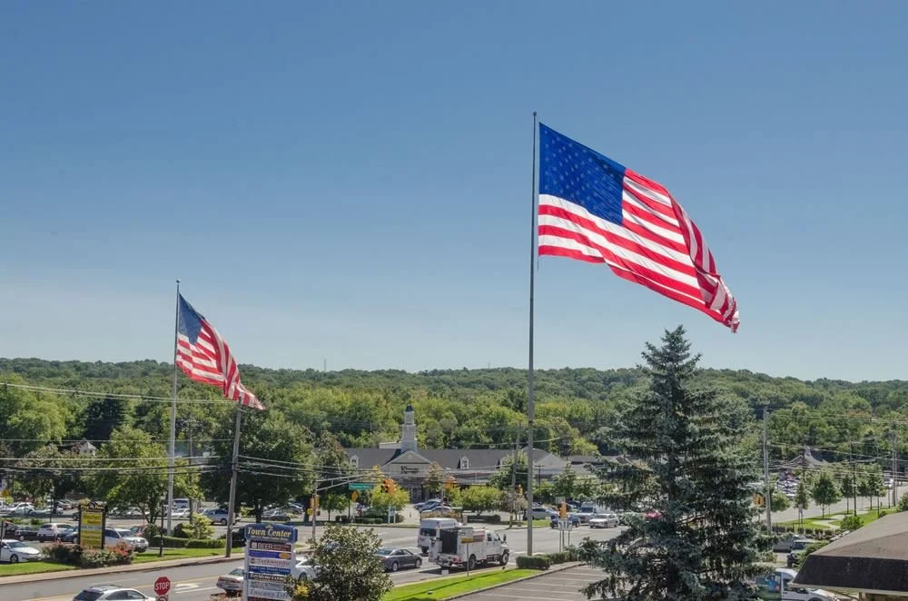Two American flags flying in a parking lot under clear blue sky, with trees and buildings in the background.