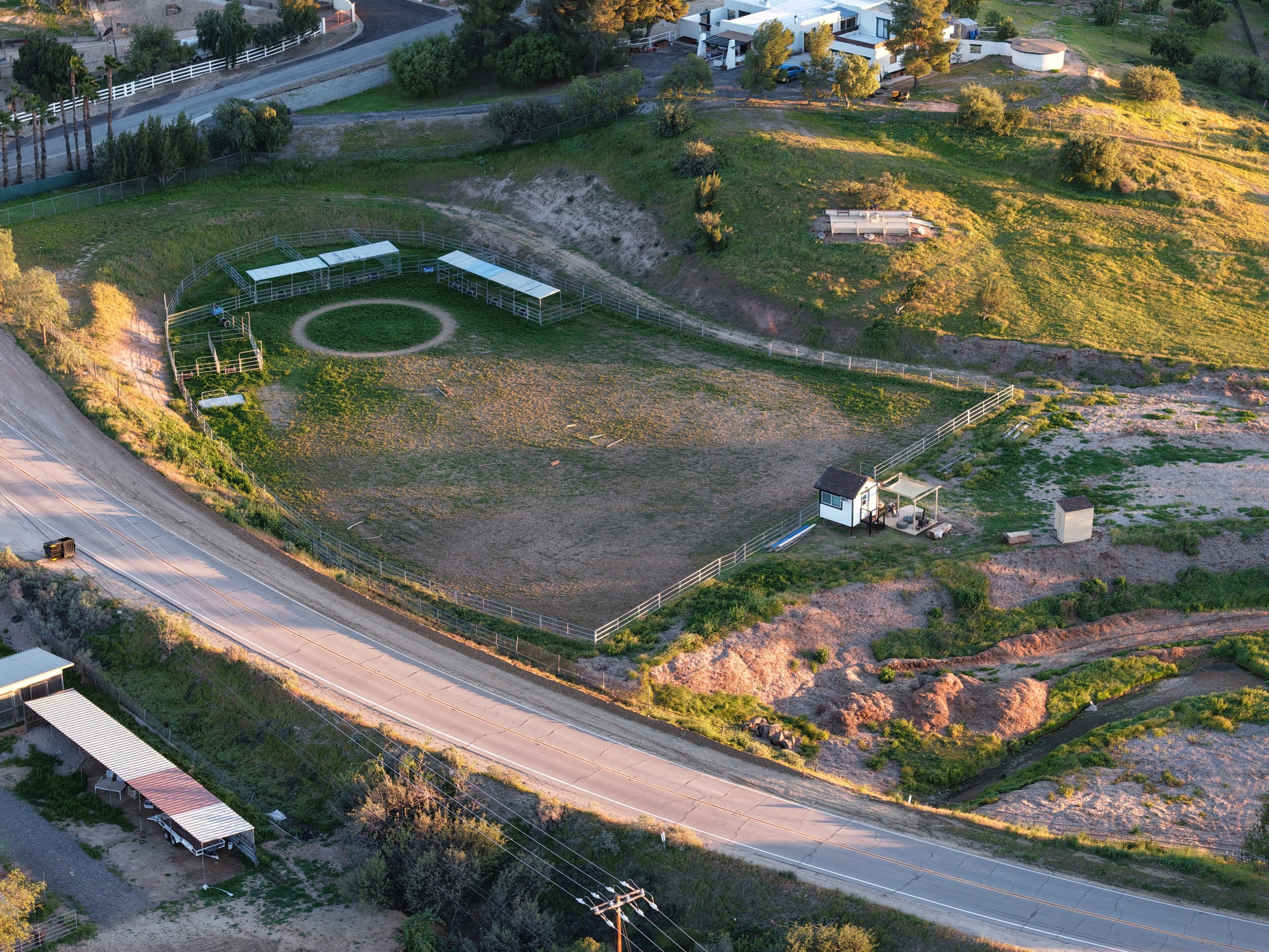 Aerial view of a fenced horse riding arena with several sections and a small structure, located beside a road with surrounding greenery and residential area.