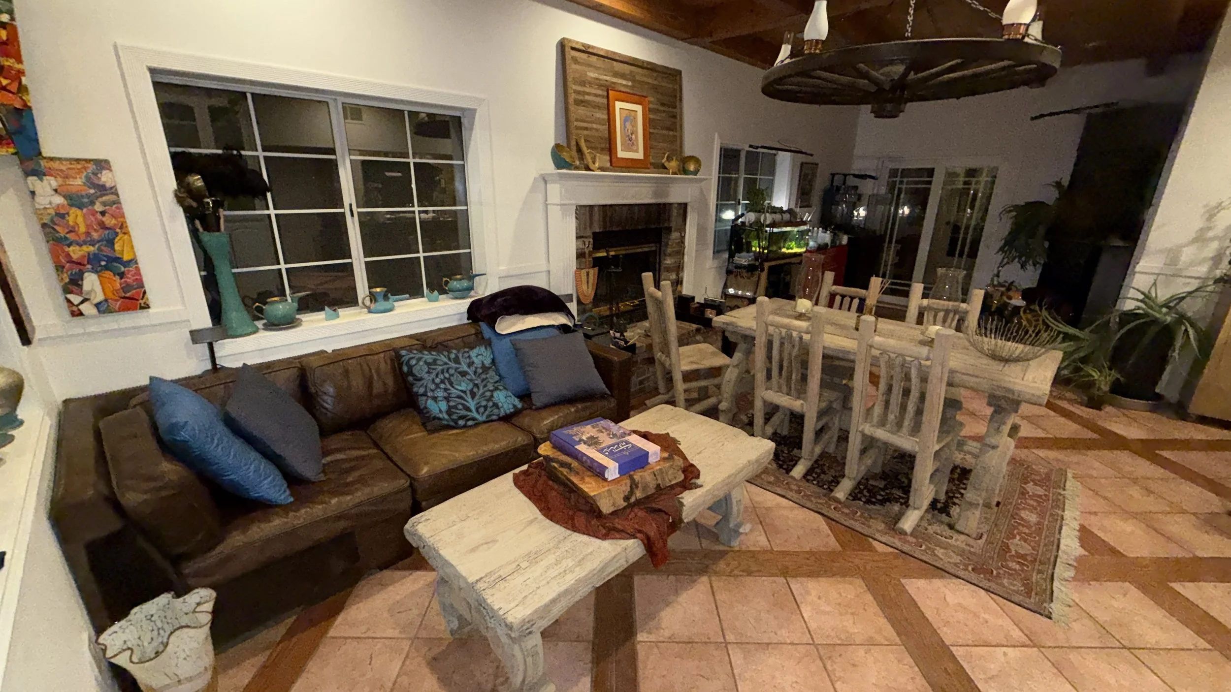 Living room with brown leather sofa decorated with blue and gray pillows, a white wooden coffee table with books, and a rustic dining table with six chairs. The room features a brick fireplace with a wooden mantel and artwork above, a large window with display of teal-colored teapots, and various plants and decorative items. The flooring is patterned tile, and a large wagon wheel chandelier hangs from the ceiling.
