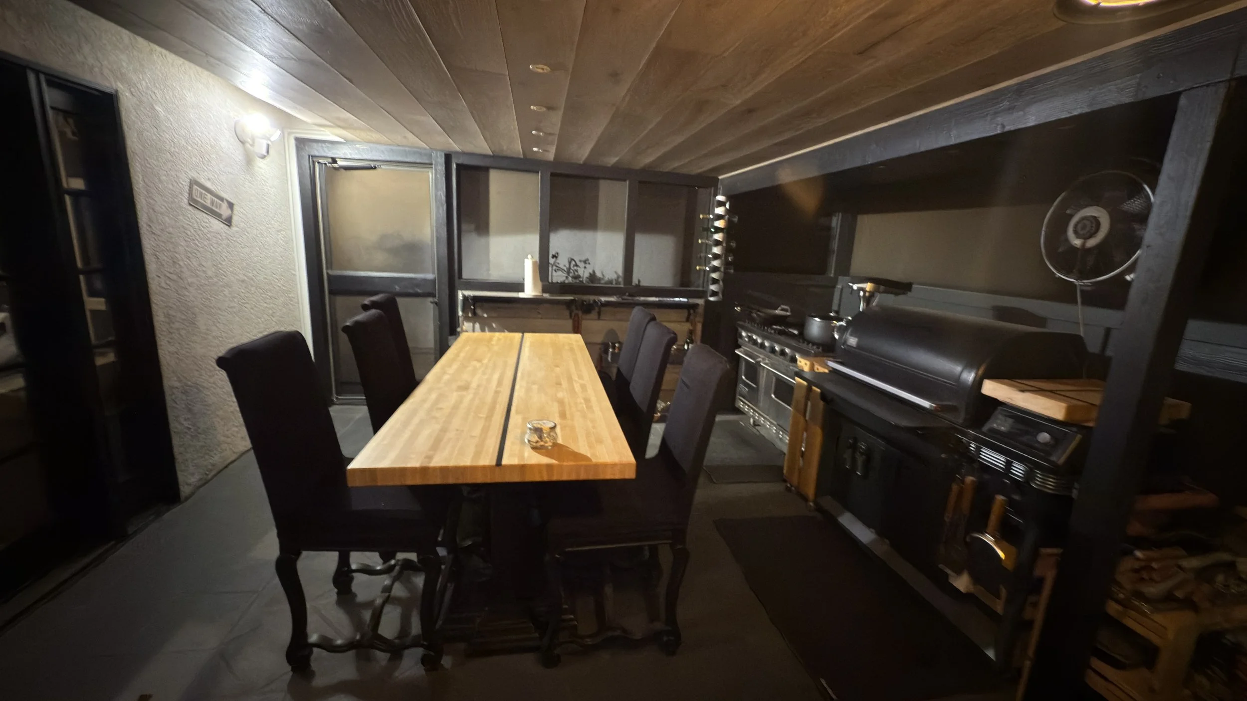 Dining area with a wooden table, six black chairs, and a black stove with pots, in a cozy kitchen with wood-paneled ceiling and dark flooring.